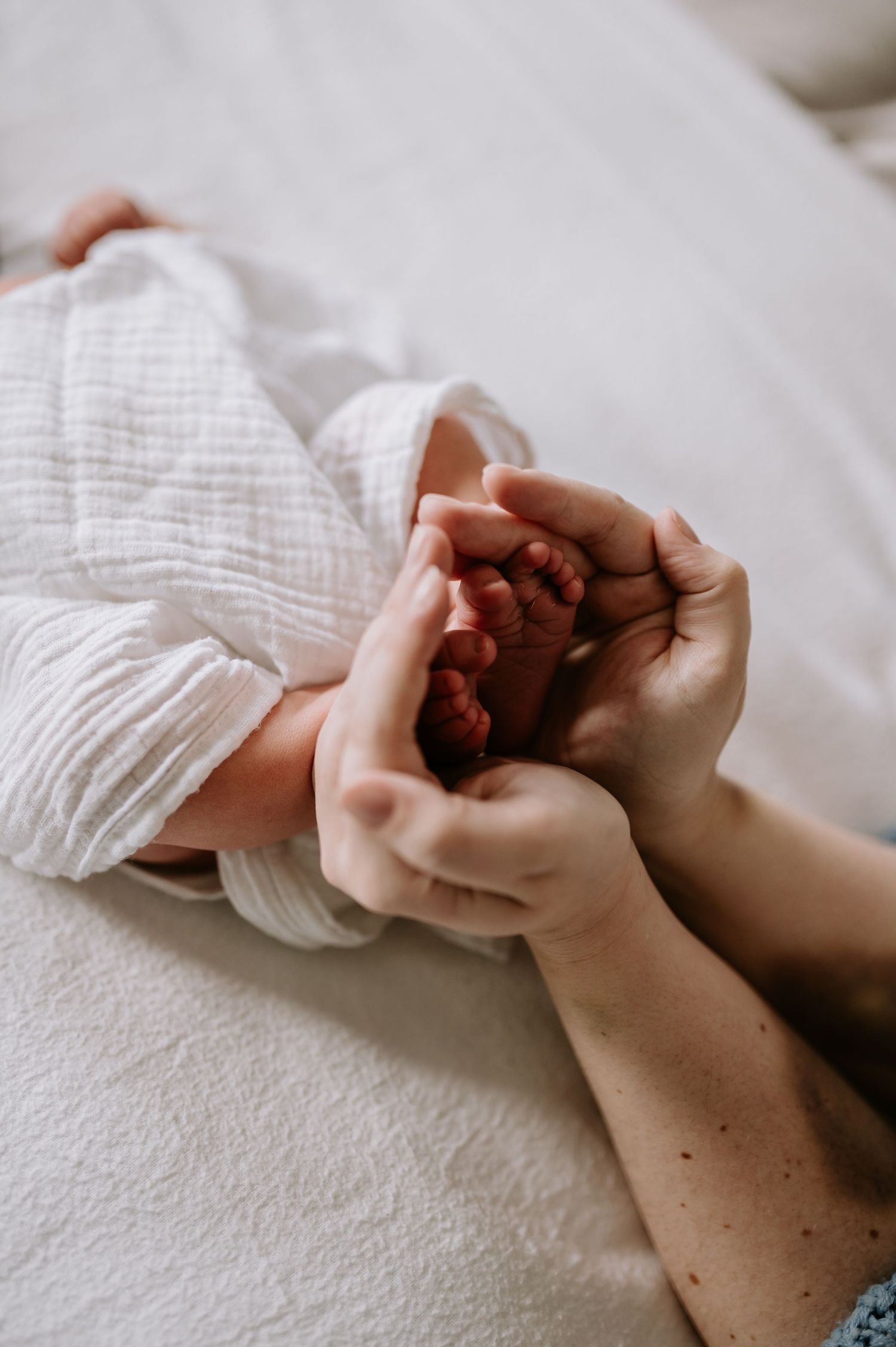 Small hands gently hold a newborn's delicate feet wrapped in soft white muslin fabric on a light surface.