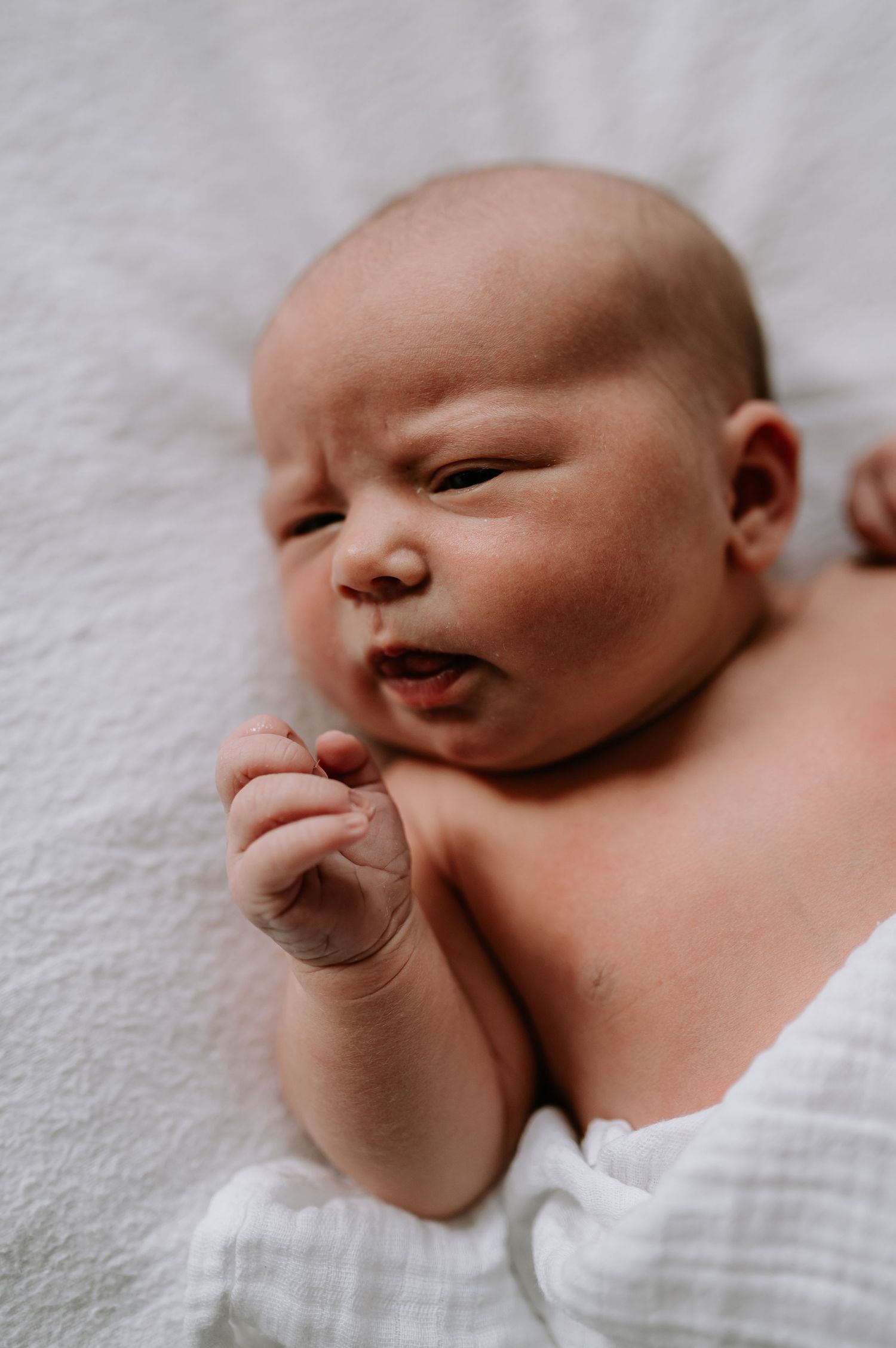 A newborn baby rests peacefully on white bedding while making a gentle expression and curling their tiny fingers.