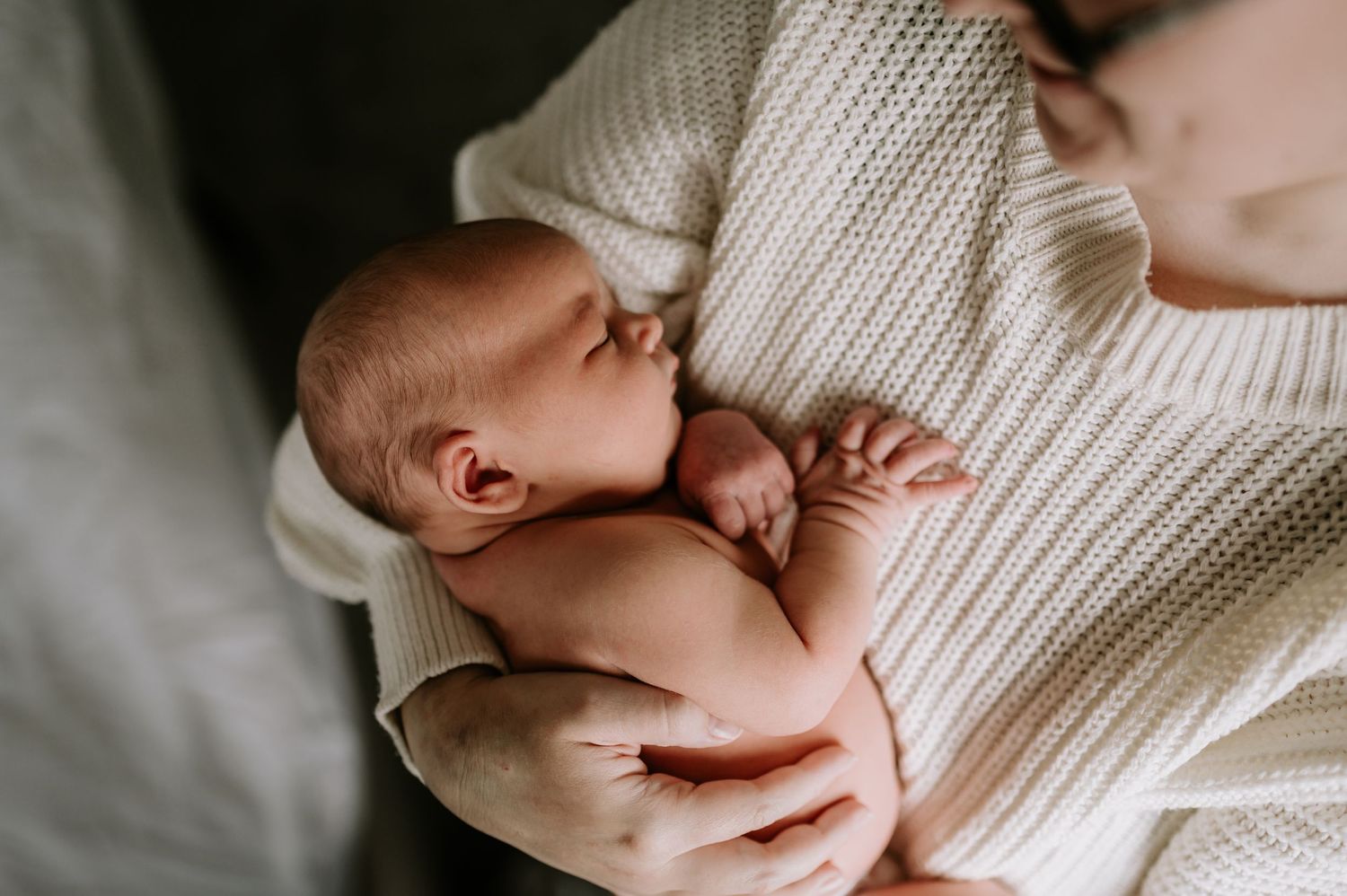 Close up of a newborn baby cradled in someone's arms wearing a white knit sweater.