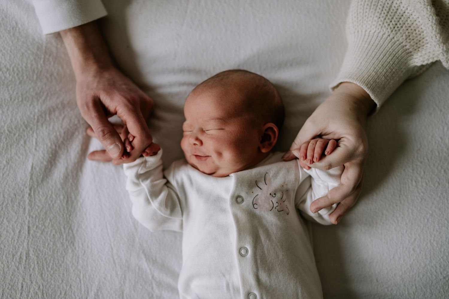 A tiny newborn dressed in white stretches on soft white fabric while gentle hands offer comfort.