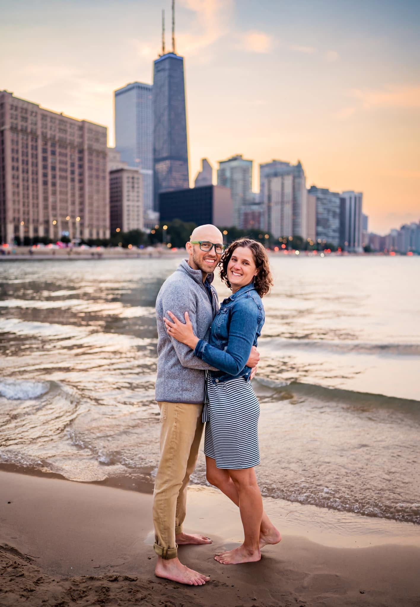 A couple embraces during sunset on a beach with the Chicago skyline featuring the John Hancock tower in the background.