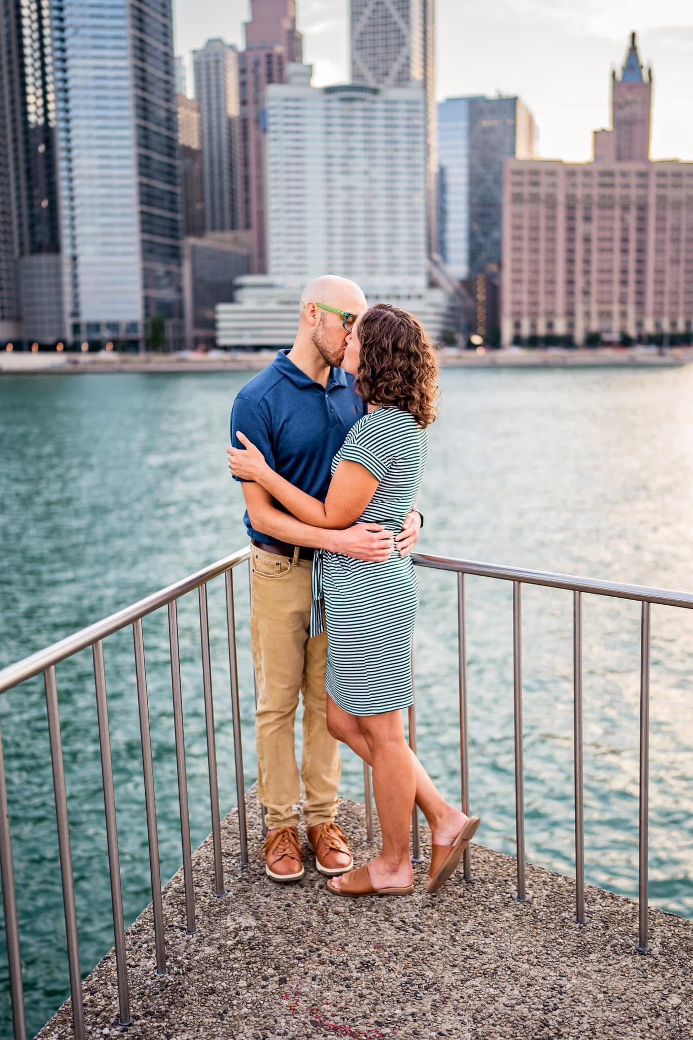 A romantic couple embraces on a waterfront overlook with the Chicago skyline and Lake Michigan in the background.