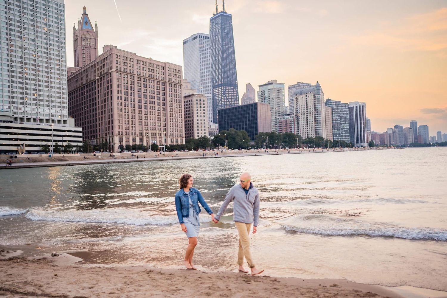 A couple walks along the Chicago River shoreline at sunset with the iconic downtown skyline in the background.
