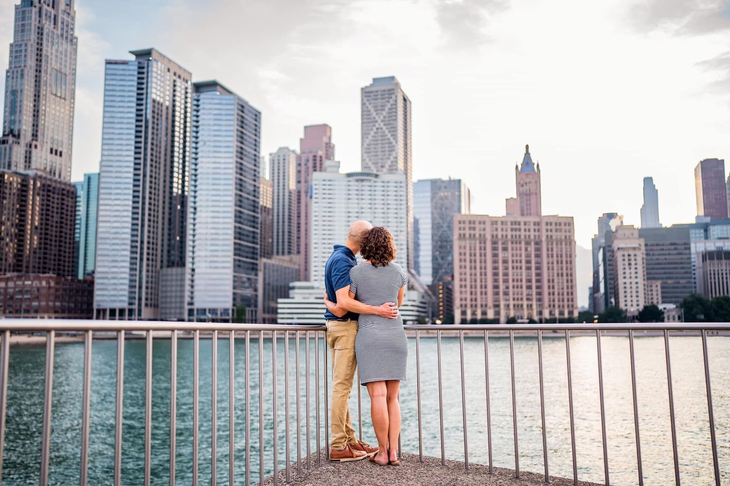 A couple embraces along the Chicago Riverwalk with the iconic downtown skyline of high-rise buildings in the background.