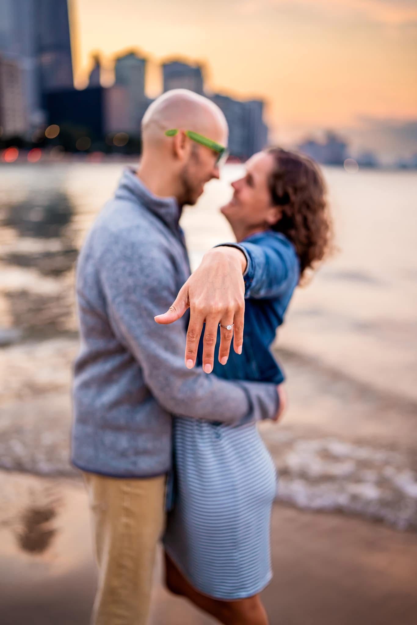 A couple showing off an engagement ring on the beach at sunset with Chicago's skyline in the background.