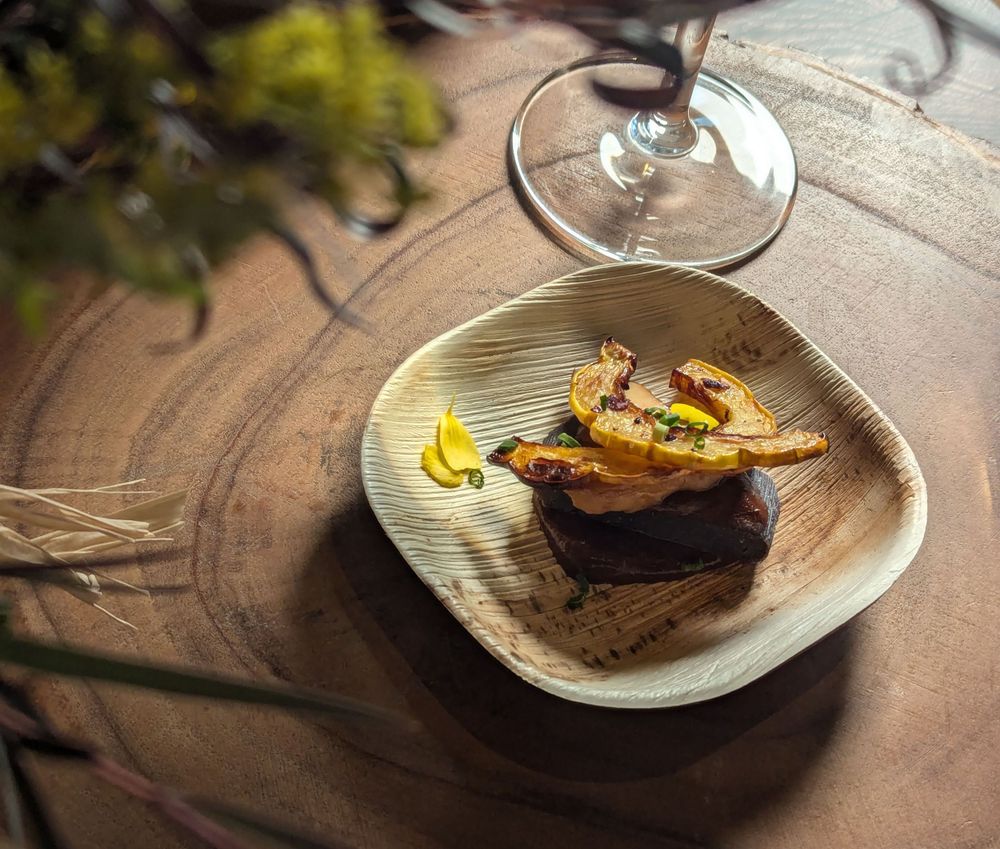 Gourmet plated dish and garnish served on a textured wood plate with wine glass in background.