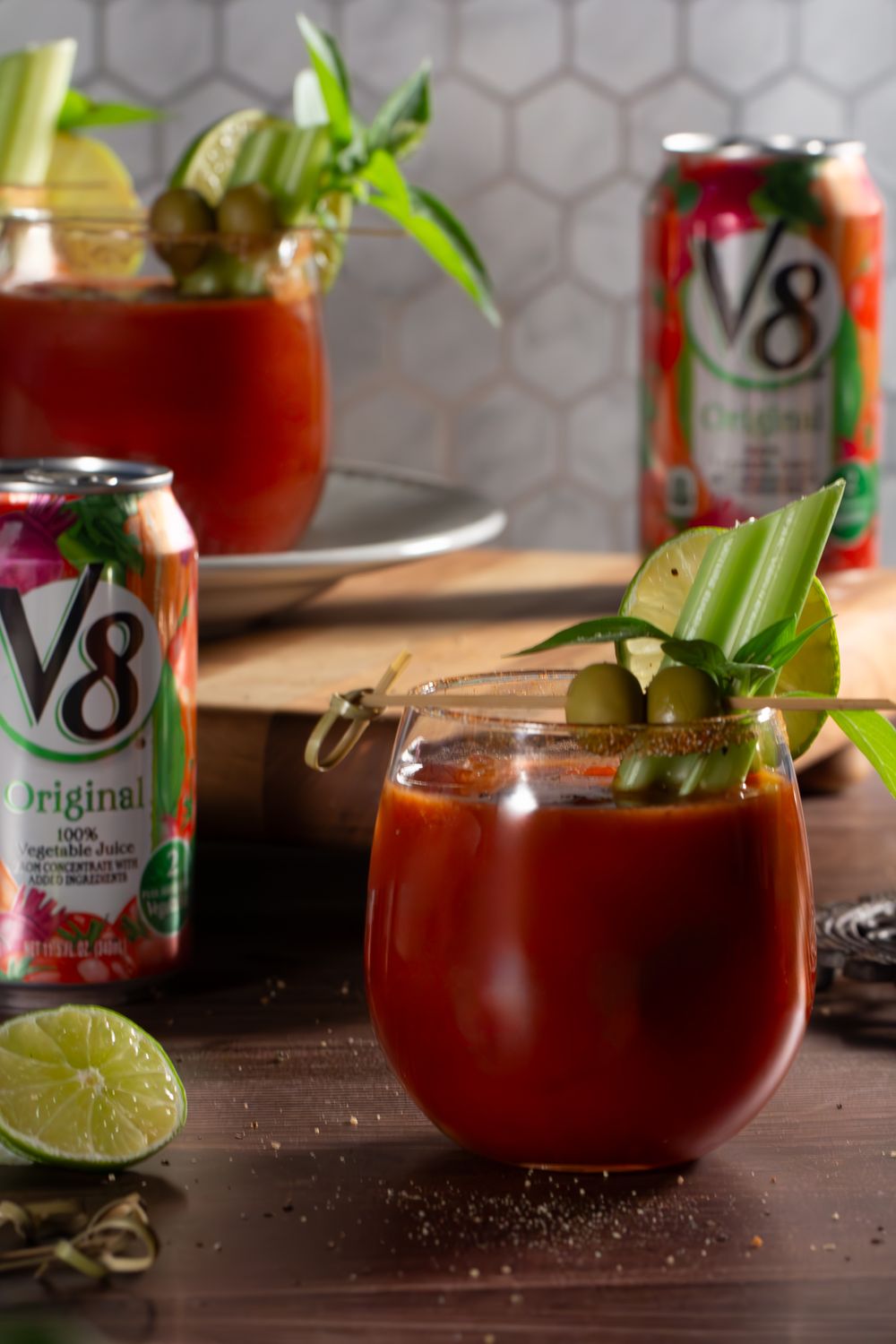 V8 vegetable juice mocktails garnished with celery, olives and lime slices alongside V8 cans on a wooden surface.