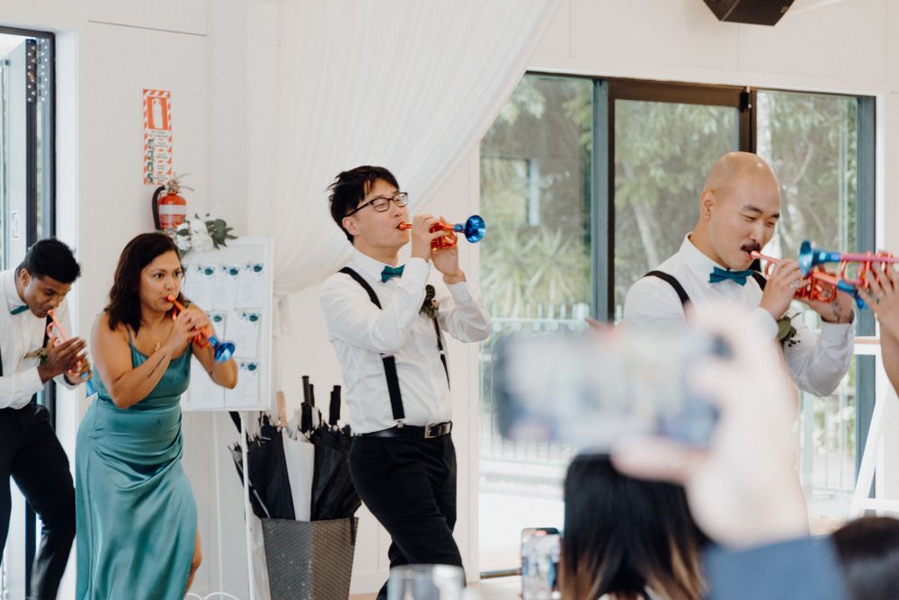 Wedding party attendees in teal dresses and black suspenders cheer captured by Apertured Media Auckland Wedding Photographer.