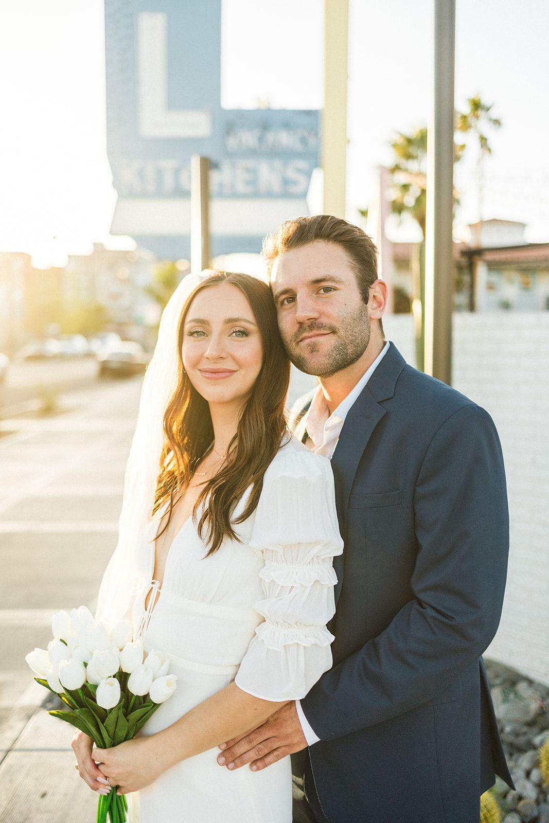 A couple in wedding attire holds white tulips while standing outside in warm sunset lighting near palm trees.