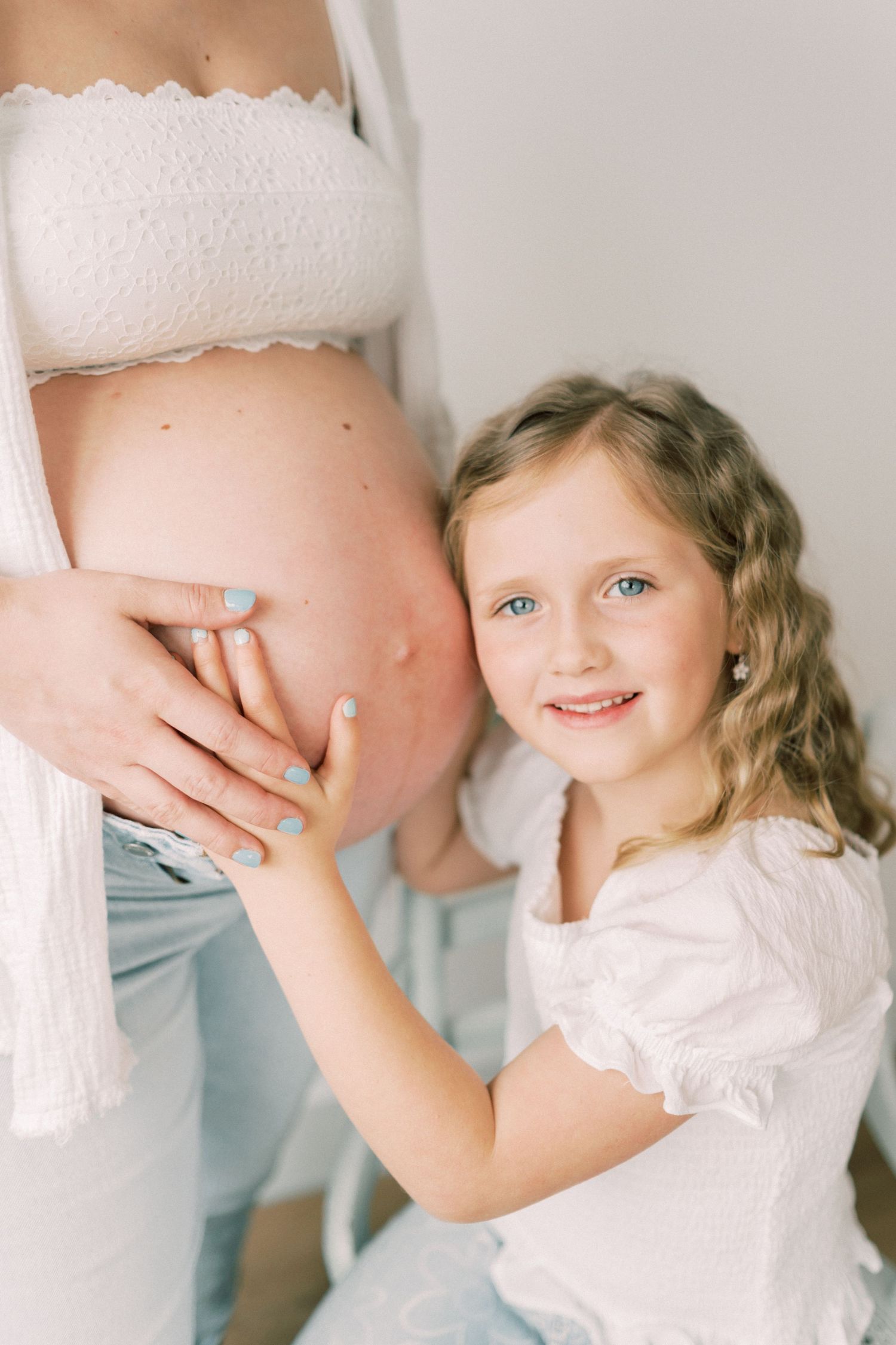 Young child in white dress touches pregnant woman's belly wearing lace bandeau top in bright indoor setting.