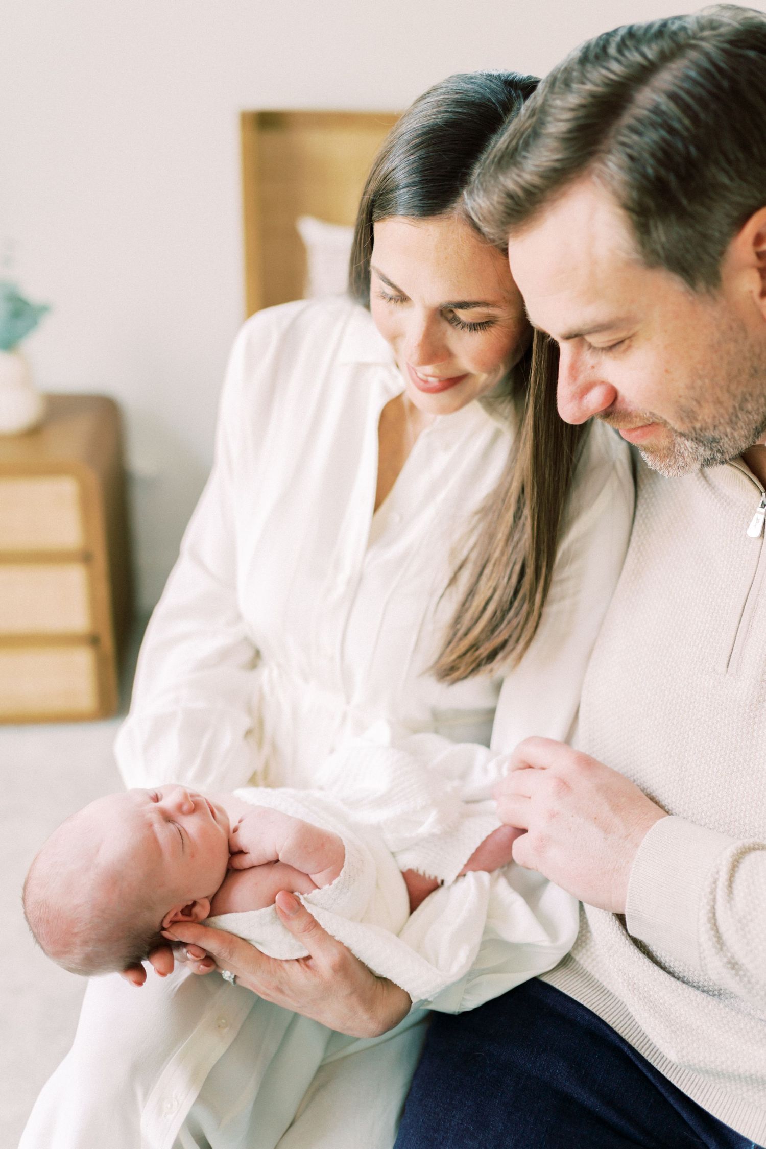 Parents in white clothing gently hold their newborn baby while sharing an intimate family moment at home.