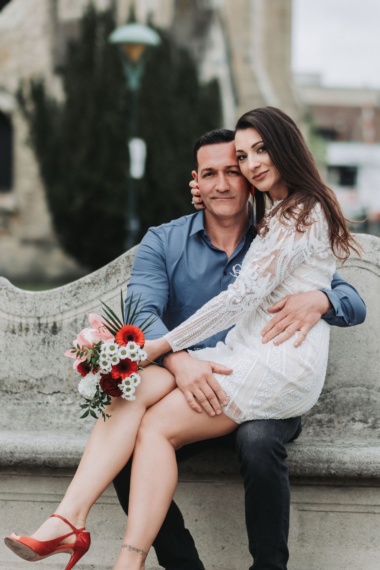 A romantic couple poses on stone steps with a small red and white bouquet against a church backdrop.