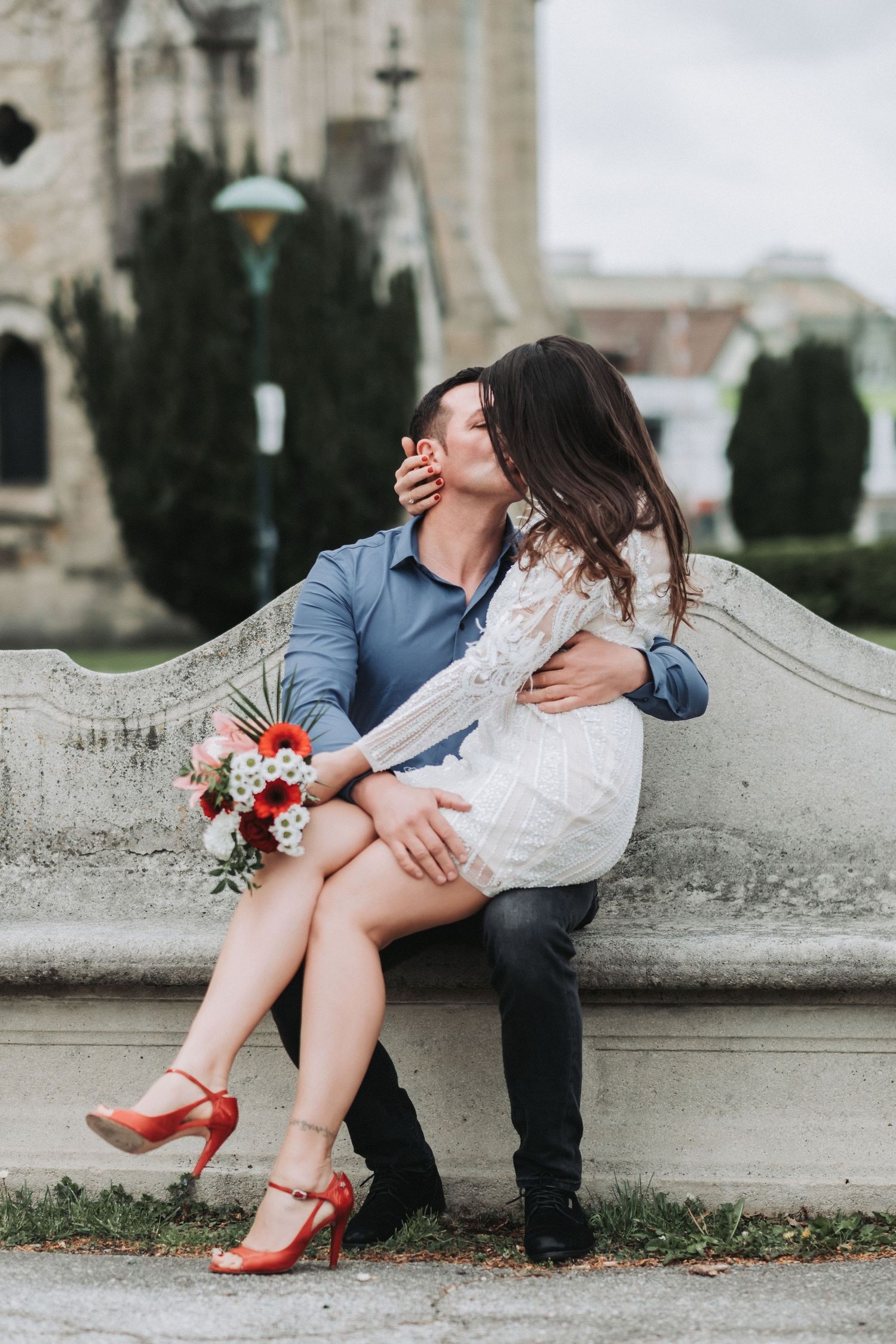 A joyful couple laughs together on stone steps with a bouquet of red and white flowers.