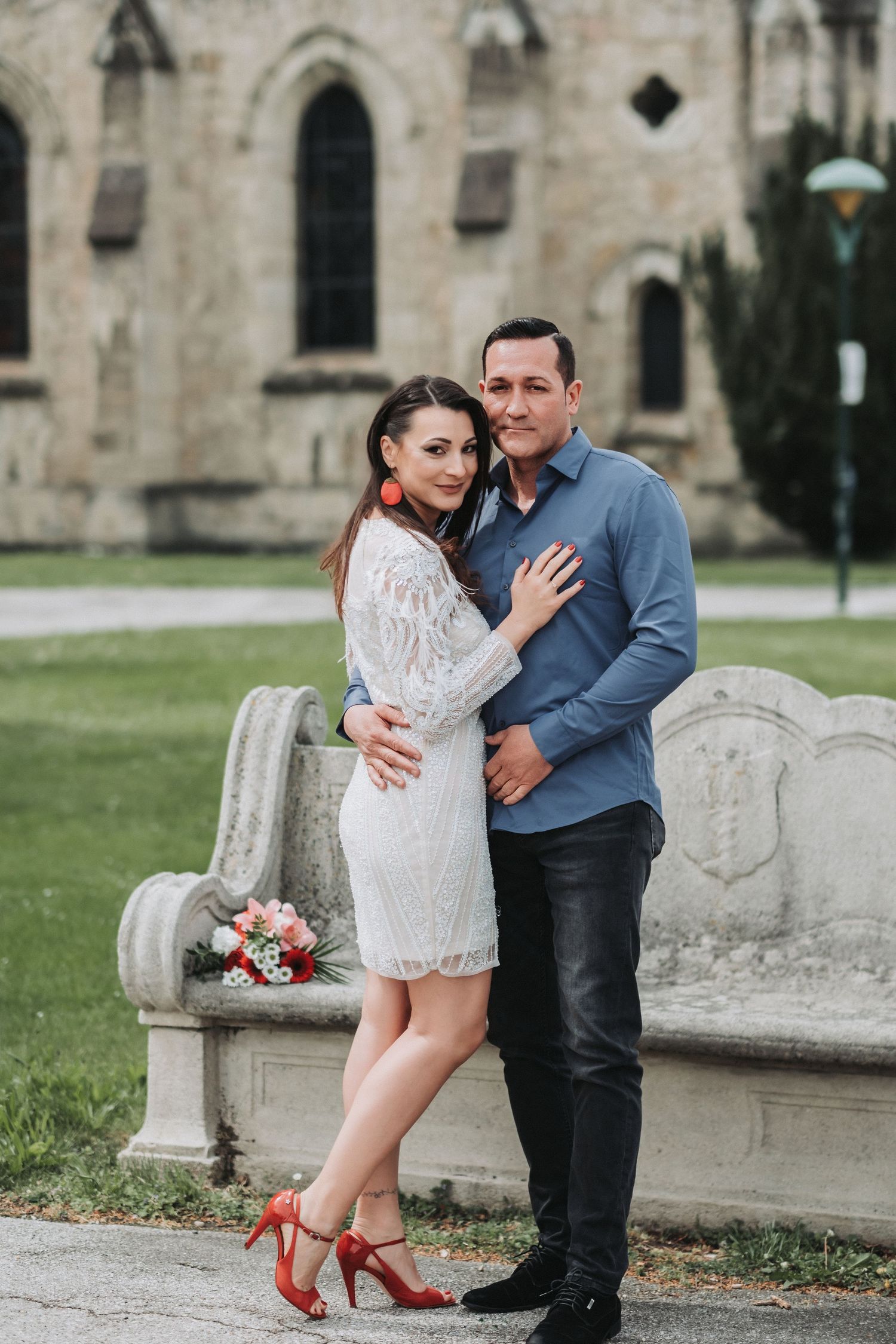 A couple poses elegantly by a stone bench with a Gothic church facade in the background.