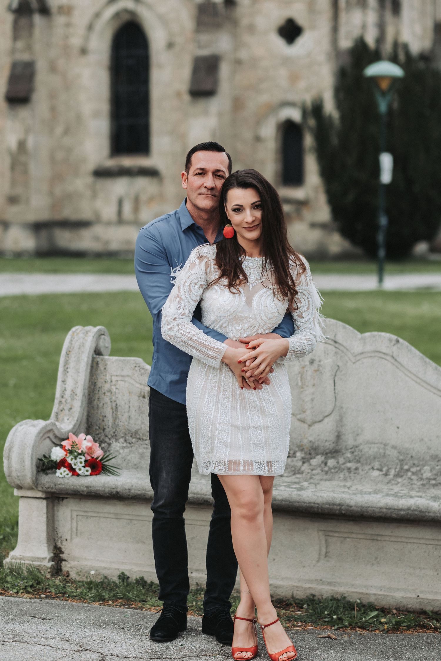 A loving embrace captured in front of stone church architecture with red and white flowers nearby.