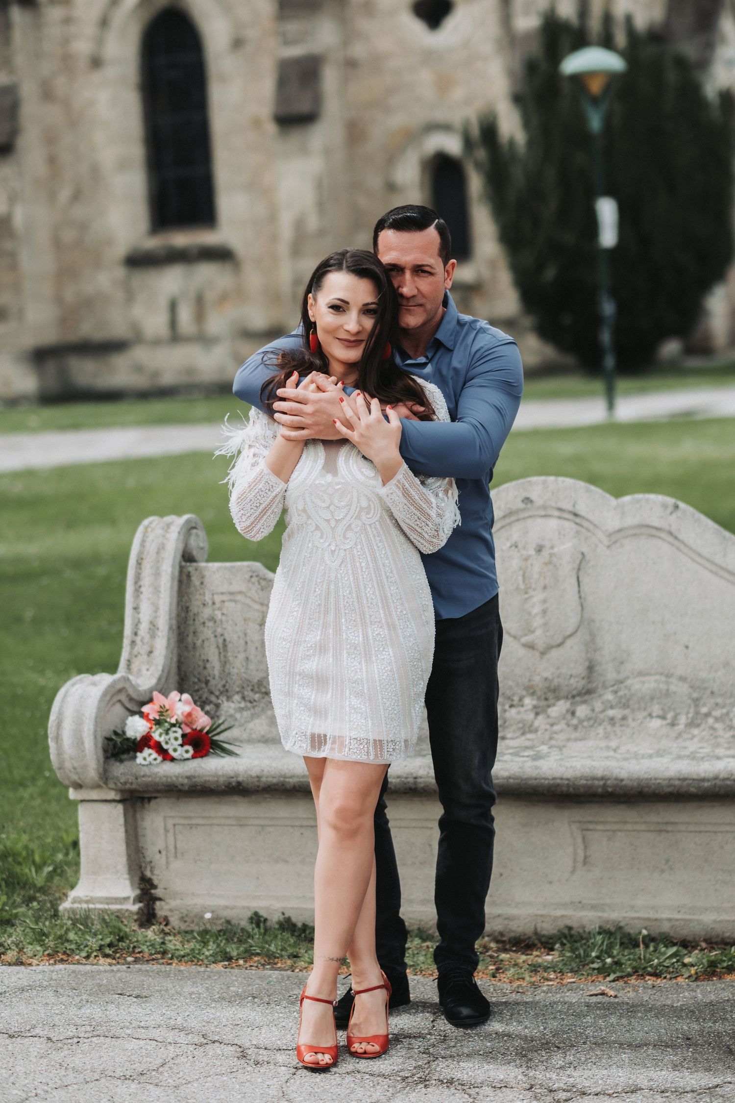 Couple embracing by a stone bench near a historic church building.