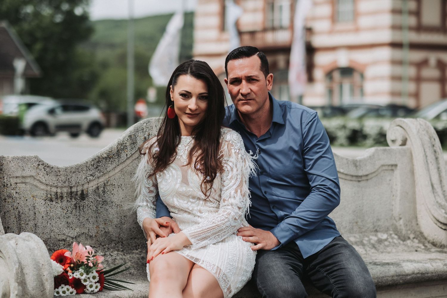 A romantic couple sitting on a stone bench, her in a white lace dress and him in a blue shirt.