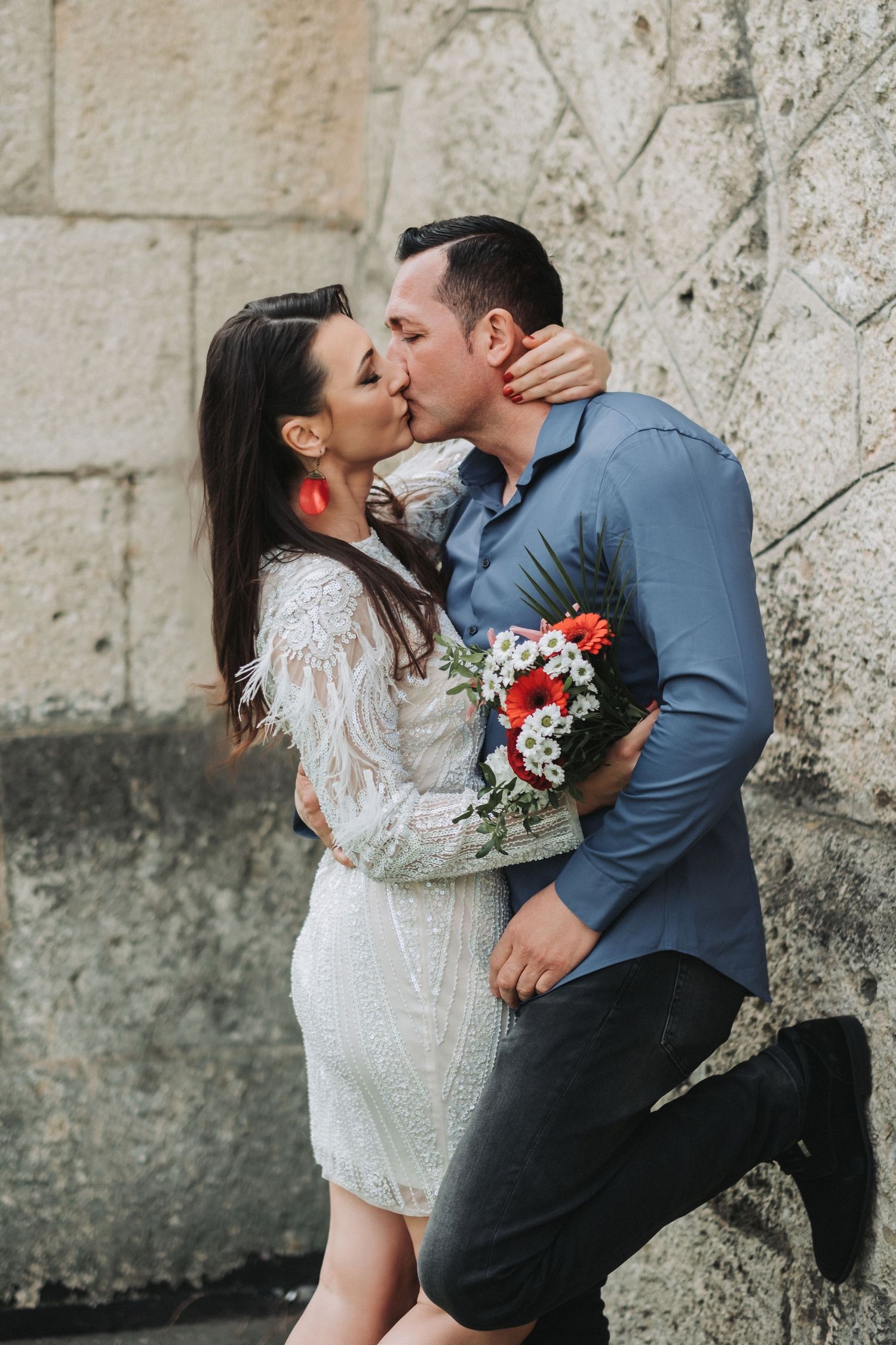 A romantic kiss captured against a stone wall backdrop with a bright floral bouquet.