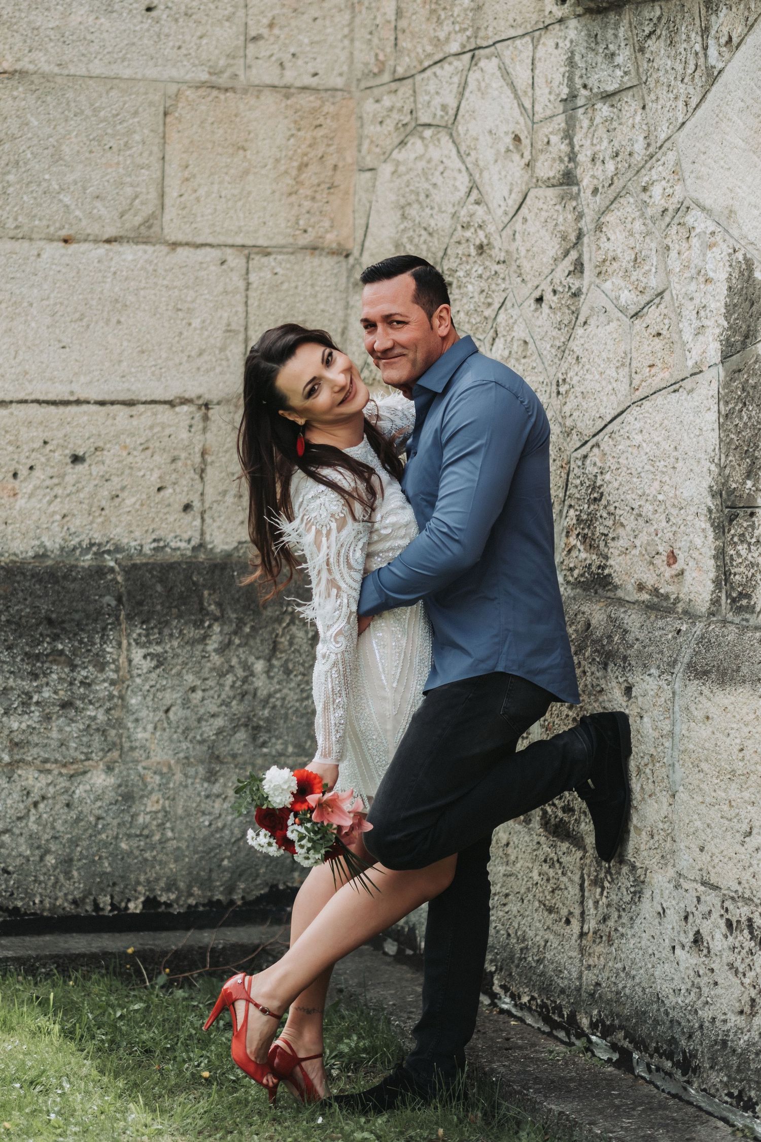 A playful couple pose against a stone wall, she holding red flowers and wearing red heels.
