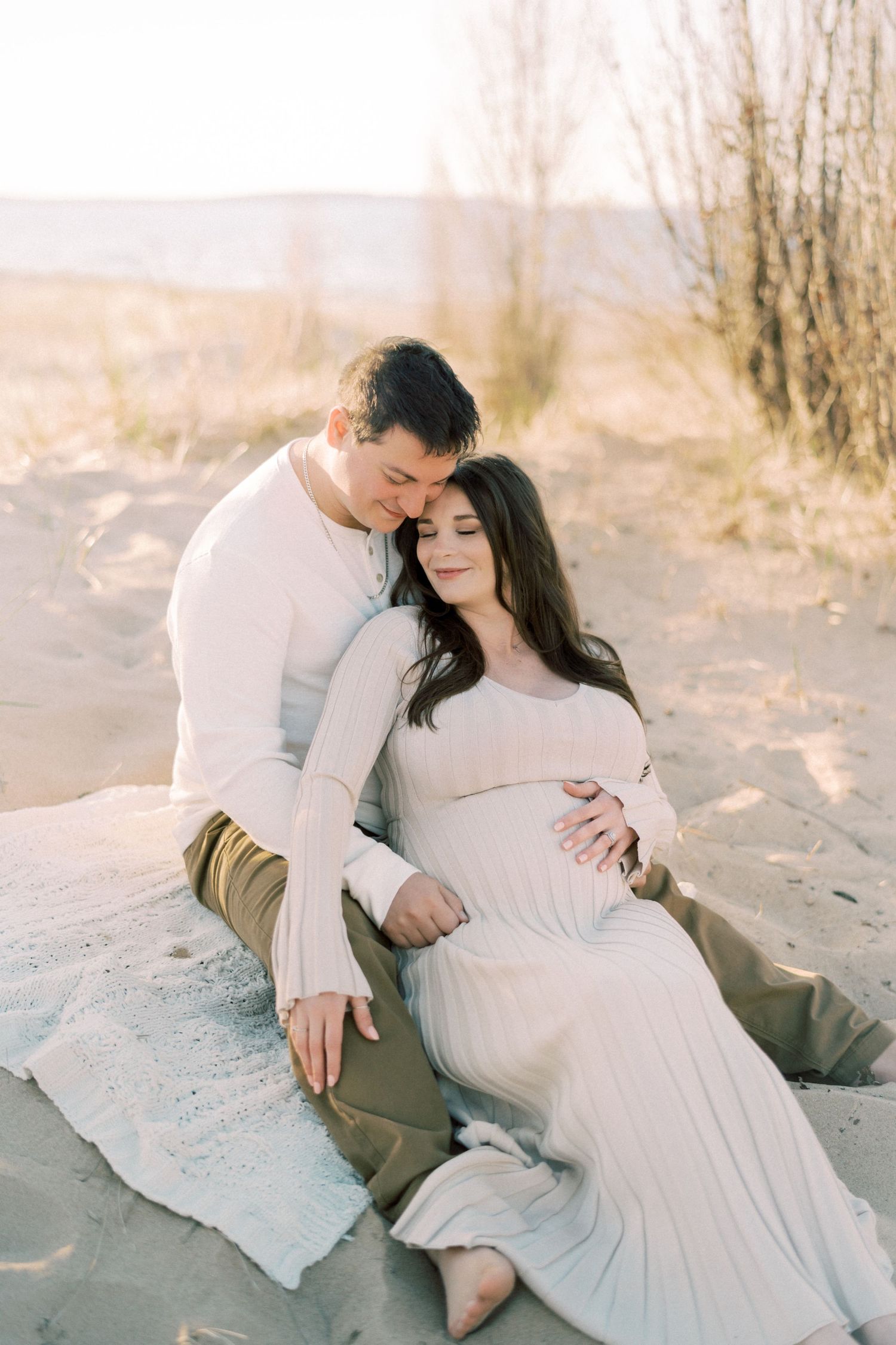 Couple sits intimately on beach blanket during sunset maternity photoshoot in dreamy natural setting.