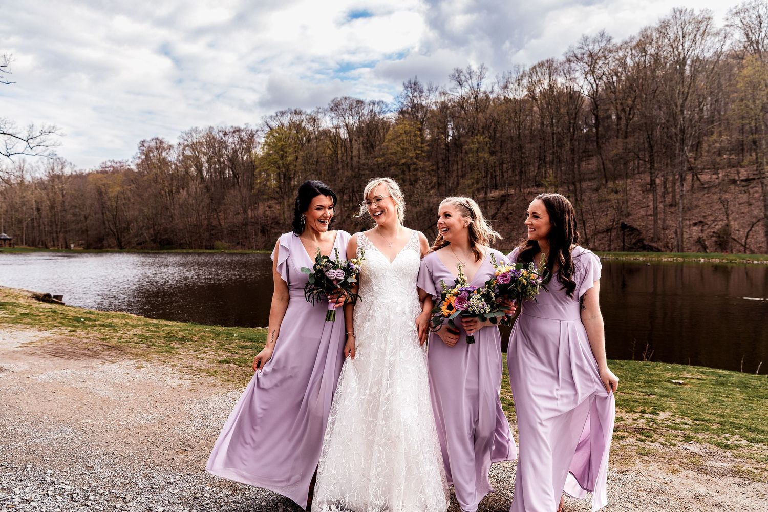 A wedding party in lavender bridesmaid dresses and a white gown hold vibrant bouquets by a serene lake in autumn.