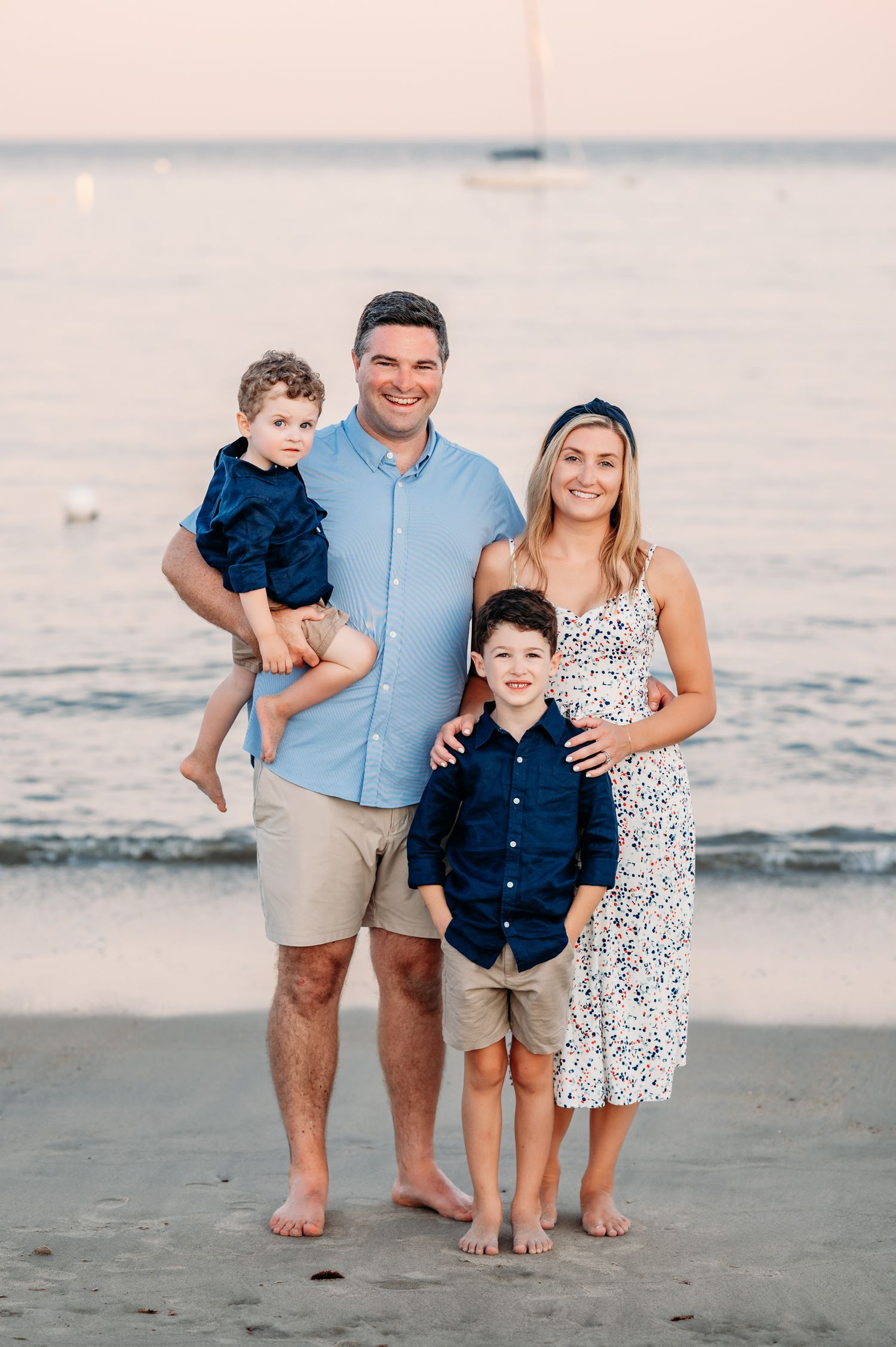 A family of four poses together on the beach at sunset wearing coordinated blue and white outfits.