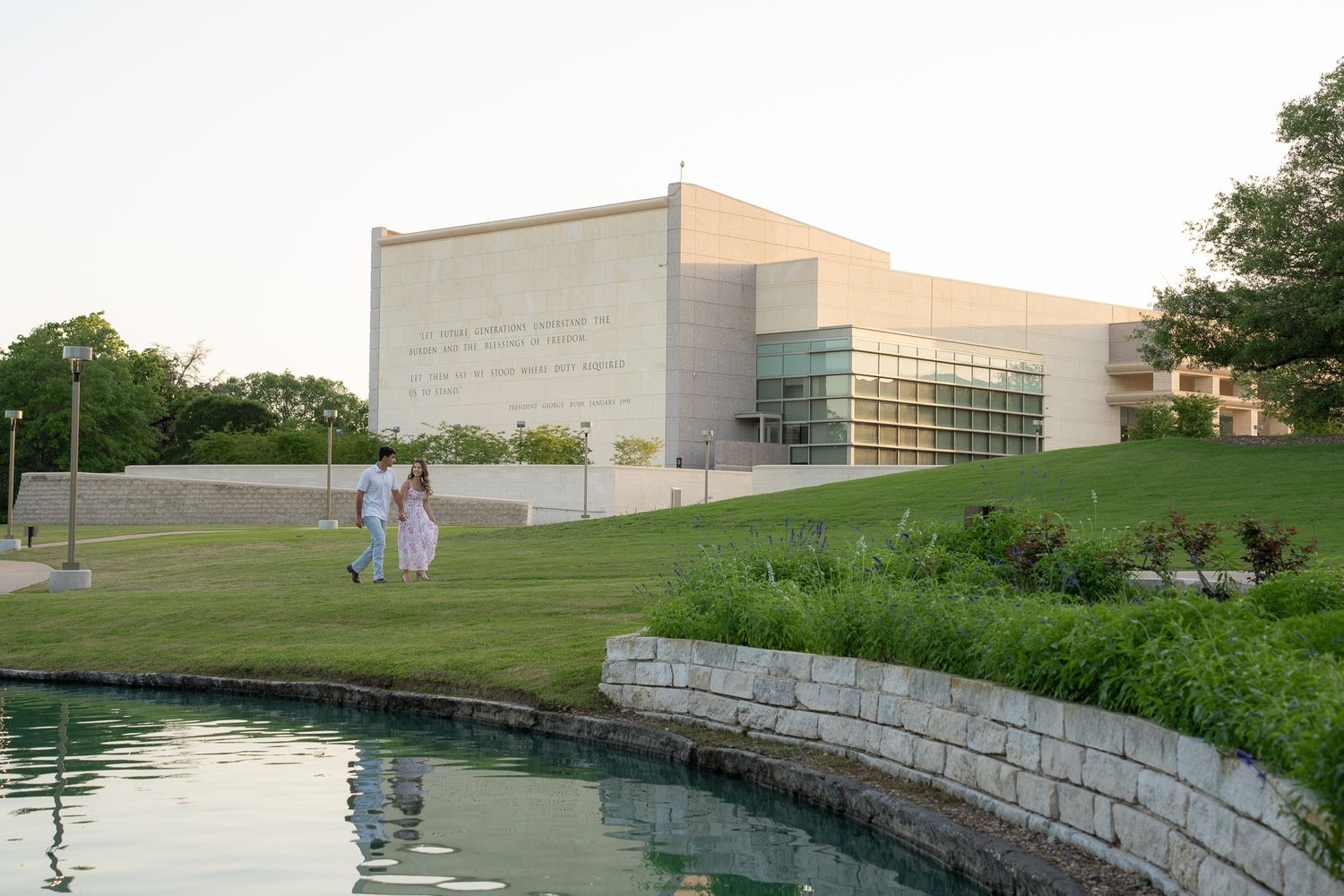 A modern institutional building with limestone walls reflects in a curved pond as people walk along the landscaped grounds.