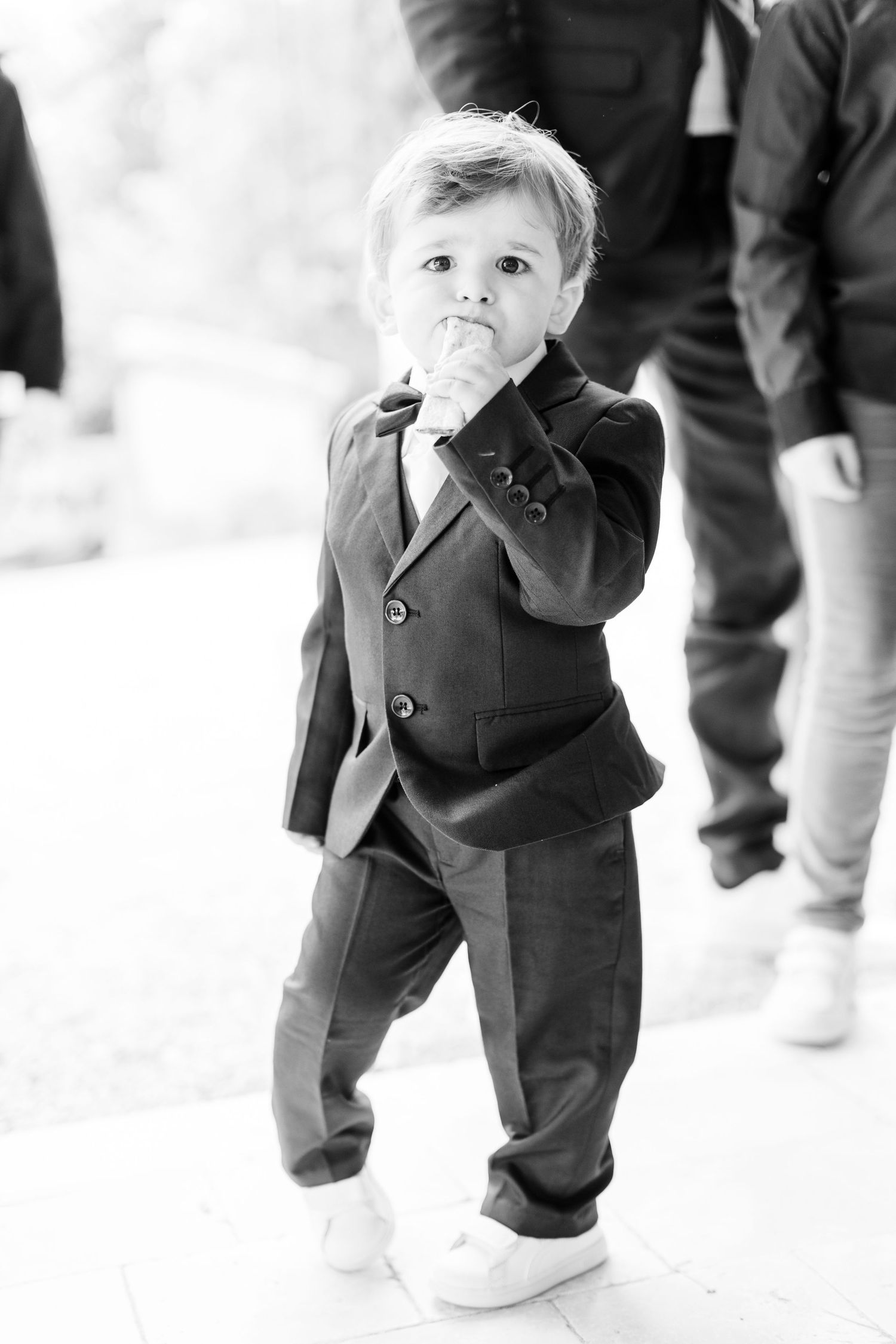 A small child in a formal suit puts their hand to their mouth while walking.
