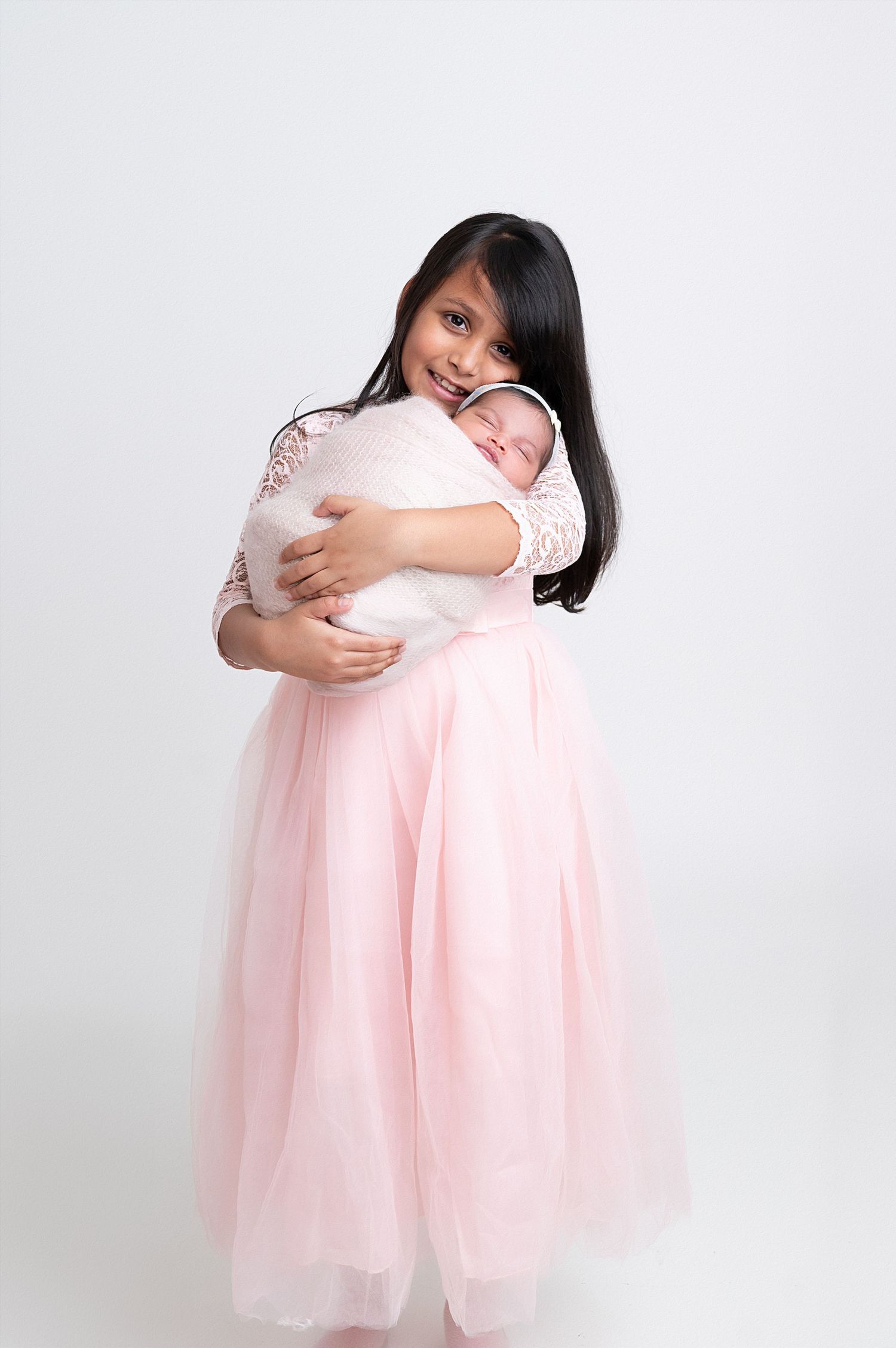 Mother in pink lace dress holding sleeping newborn against white background.