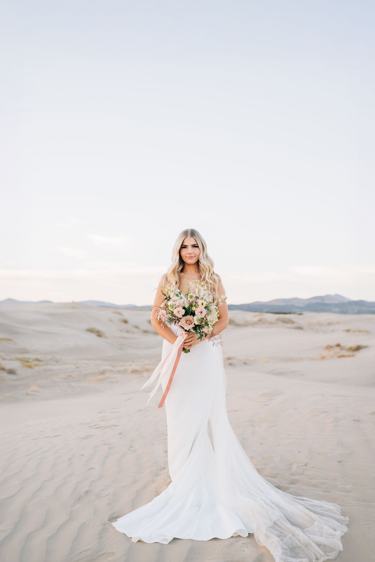 A bride in a flowing white dress holds a bouquet while standing gracefully on sand dunes at sunset.