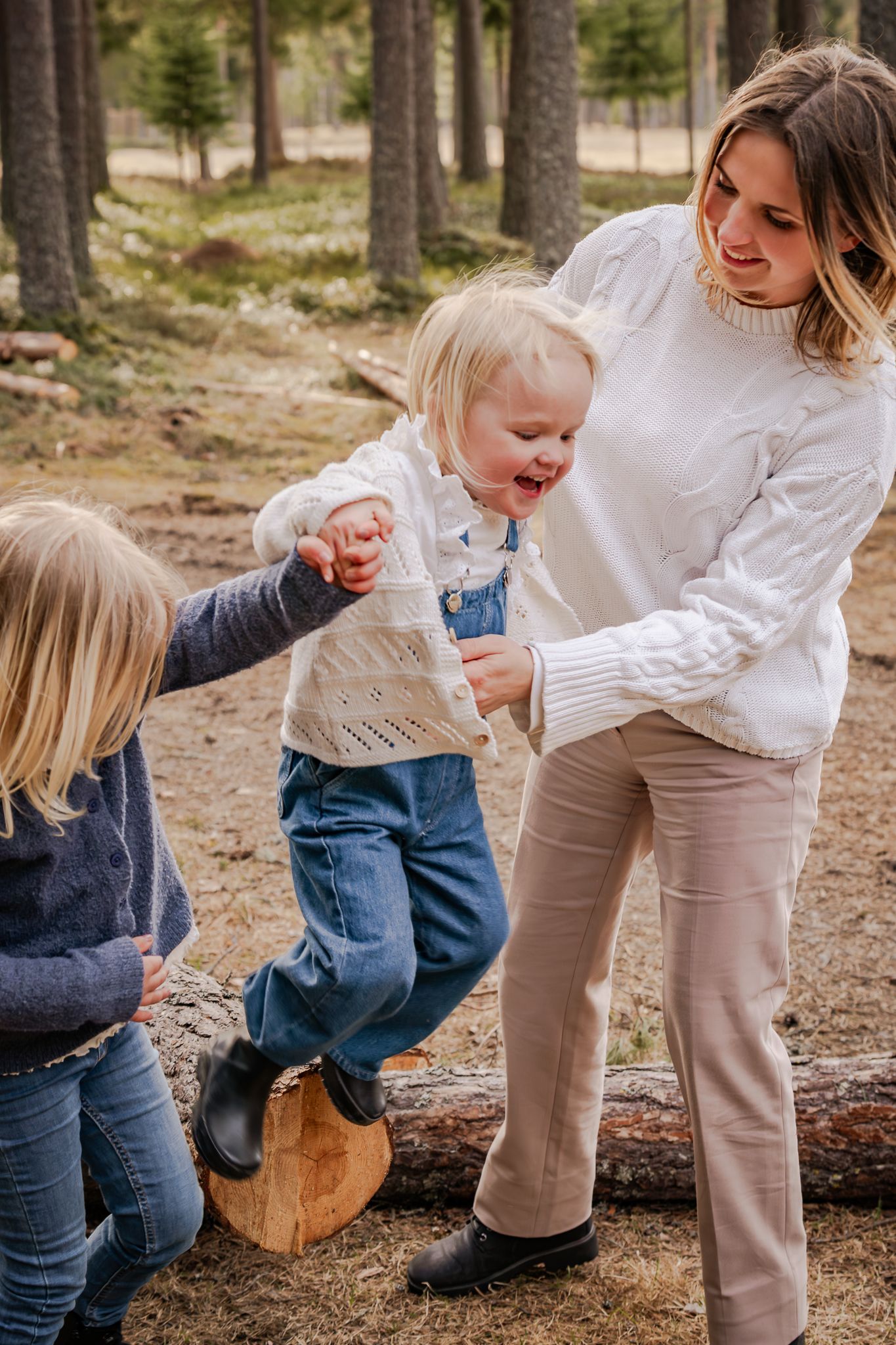 Lekfull och glädjefylld bild på mamma som håller i döttrarna som hoppar ner från en trästock i skogen. Fotograf, familjefotograf, Umeå