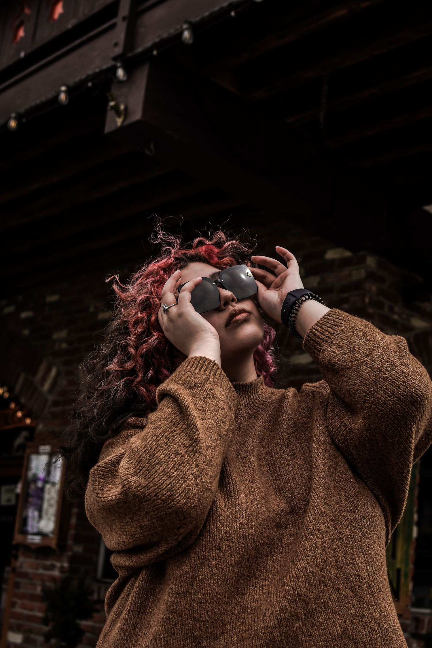 Person in brown sweater with pink hair wearing sunglasses against dark background in artistic portrait.