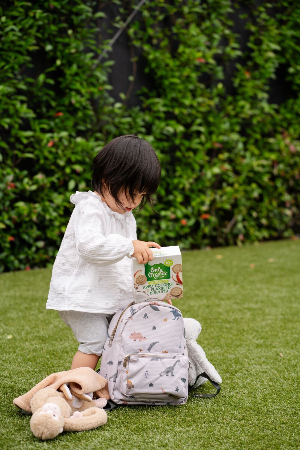 Child sits on grass examining Only Organic snack pack next to backpack and plush toys.