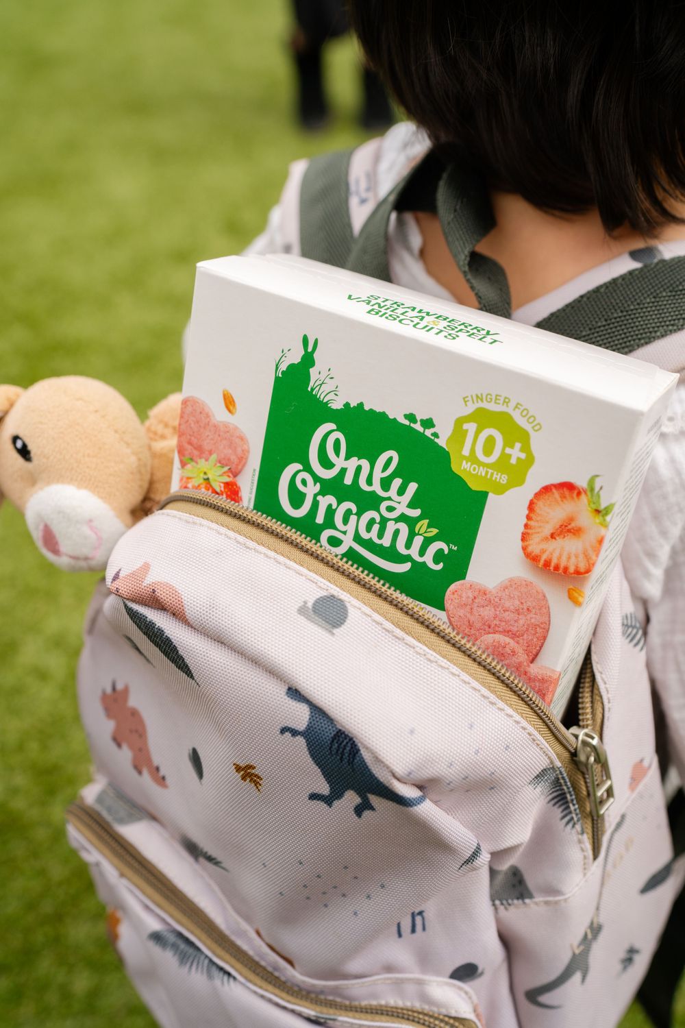 Close-up of Only Organic snack box attached to patterned backpack with plush toy.