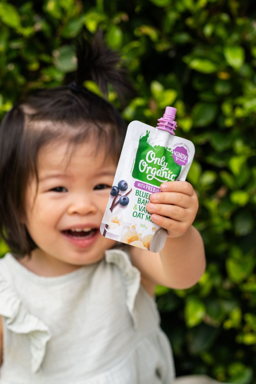 Child holds up Only Organic blueberry snack pouch while smiling outdoors.