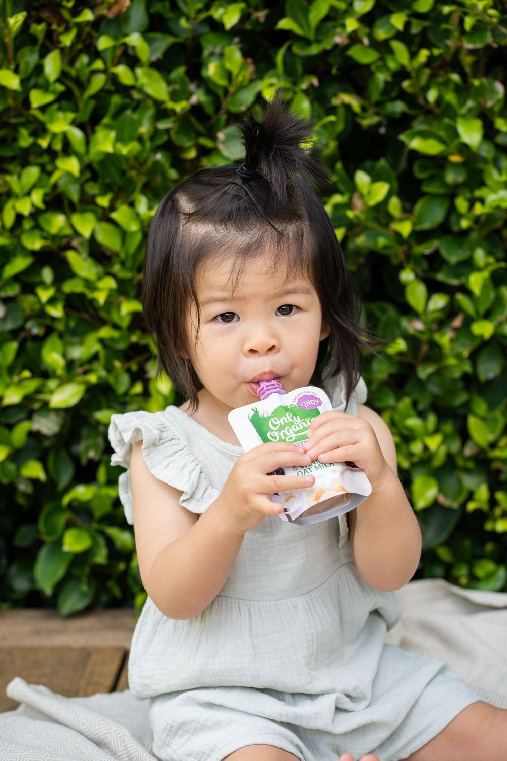 Child in white dress enjoys organic snack pouch against green hedge background.