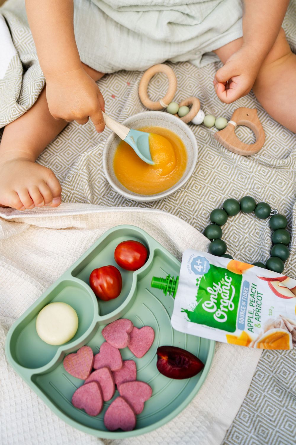 Healthy snack spread with organic pouches, tomatoes, and heart-shaped treats on gray blanket.