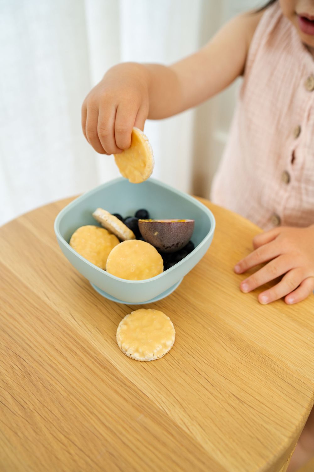 Small hand reaches for cookie from blue bowl containing snacks on wooden table.