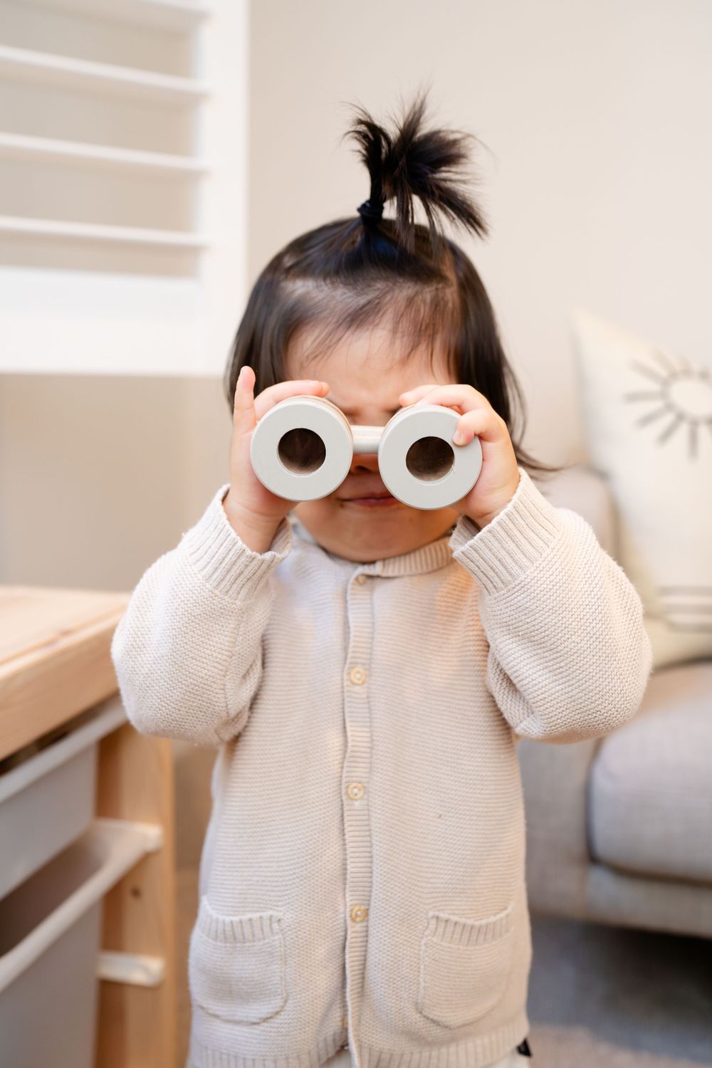 Child wearing cream cardigan holds up toy glasses in playful pose at home.