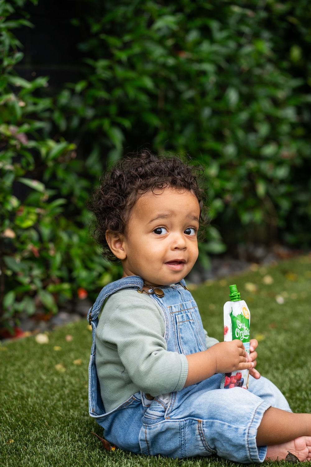 Small child in denim overalls sits on grass holding an organic snack pouch.