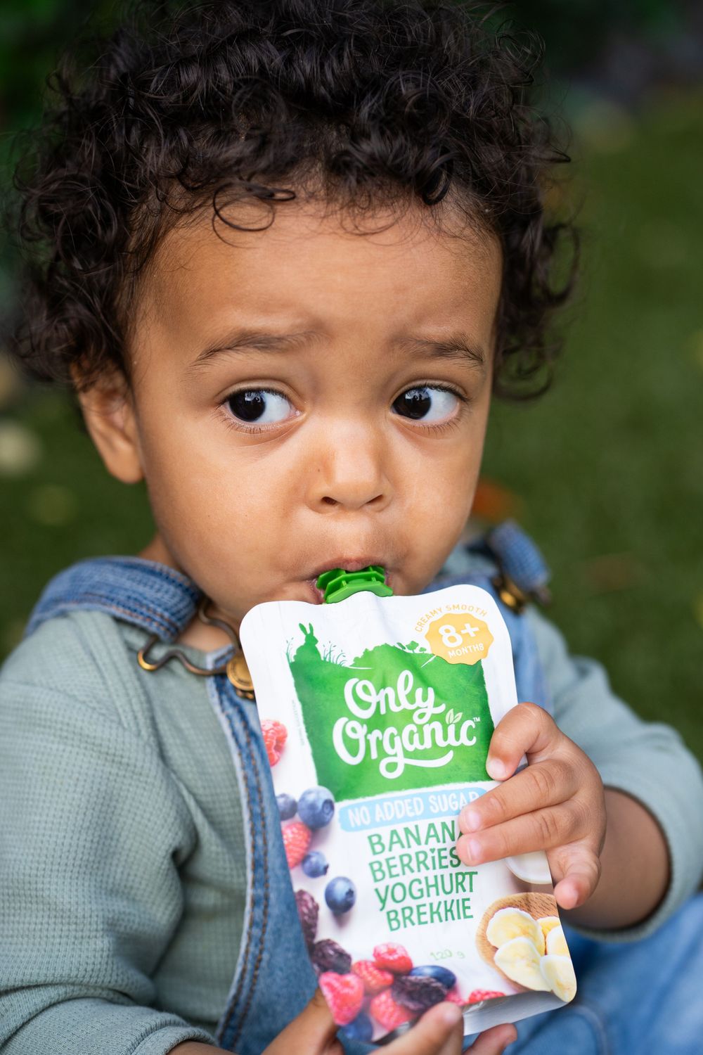 Young toddler in denim overalls enjoys an organic yogurt smoothie pouch while sitting outdoors.
