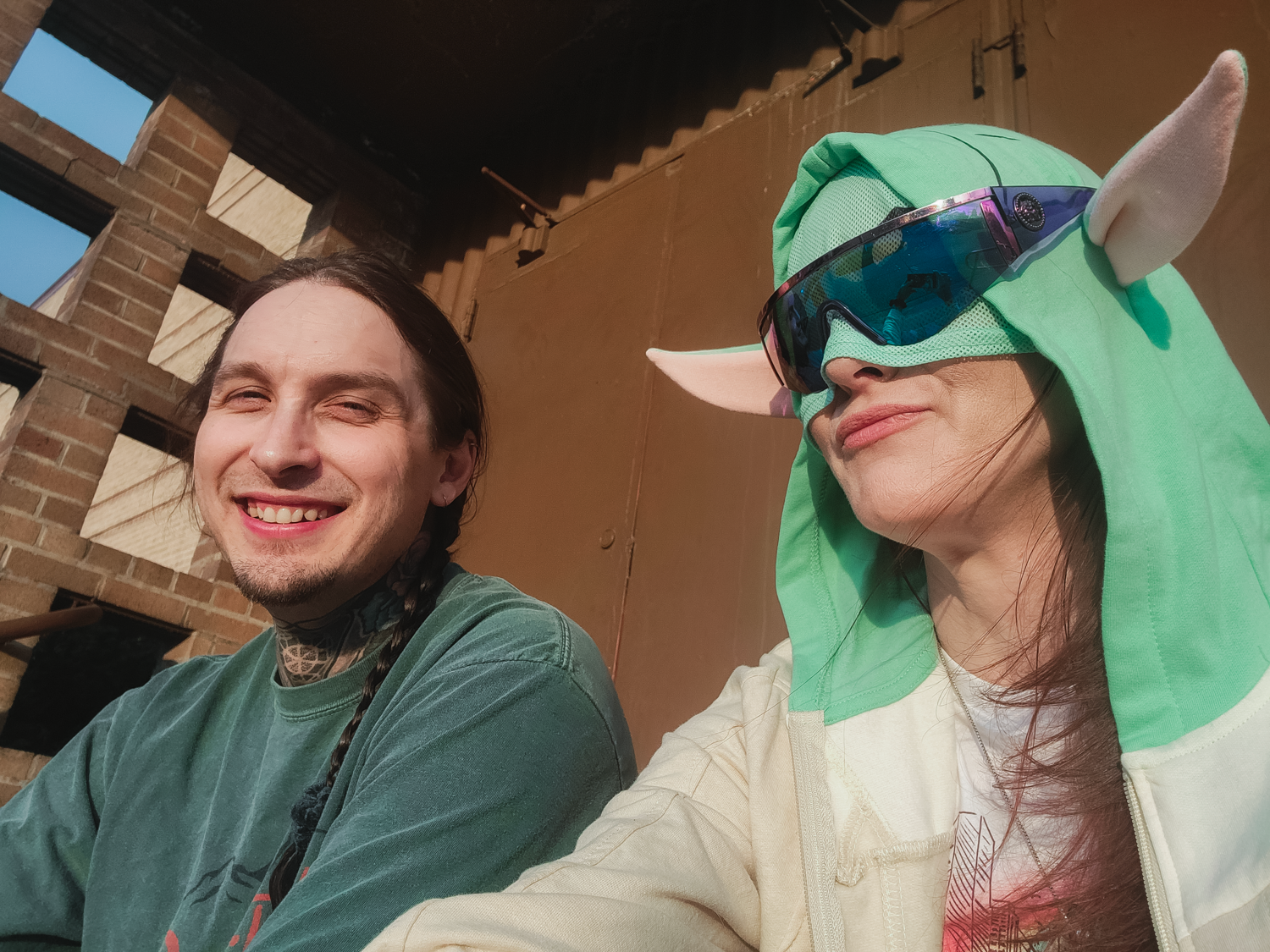 Two friends in casual wear taking selfie, one wearing green unicorn hood costume.