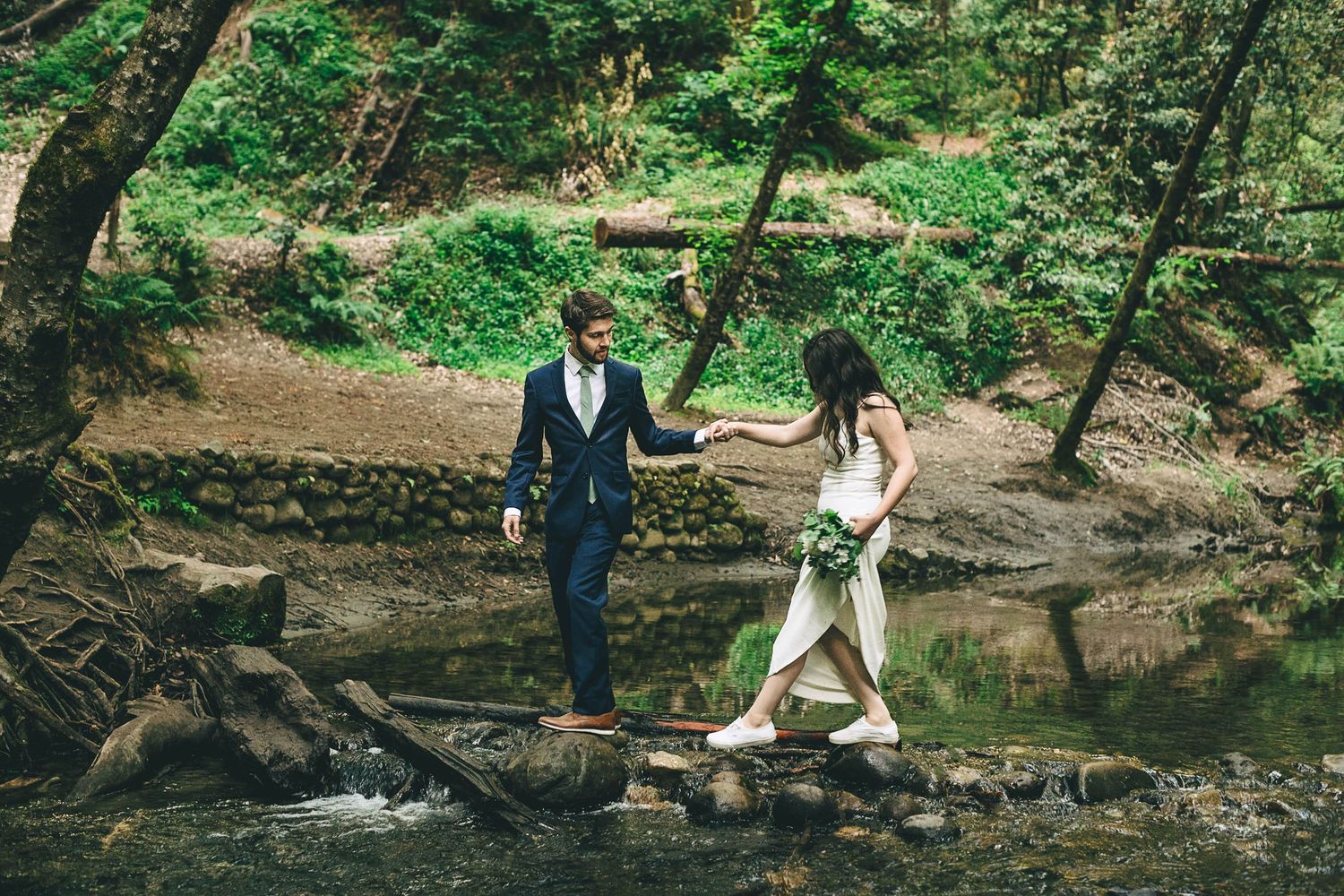A couple crosses a creek in a lush forest setting during a romantic nature photoshoot.