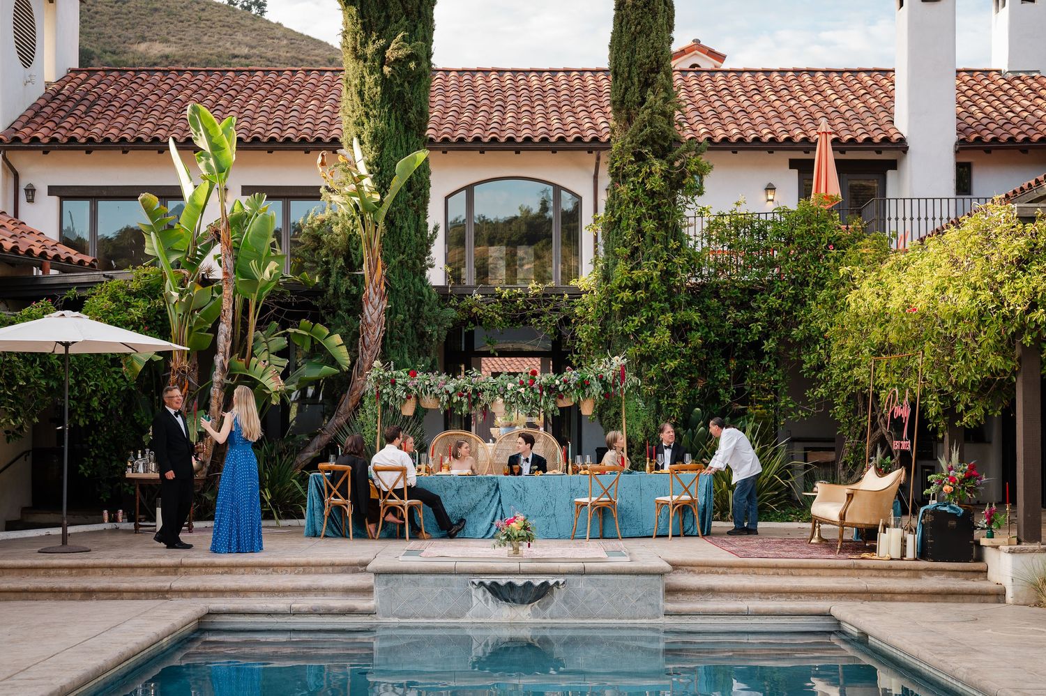 Elegant outdoor poolside gathering with people seated at a long table beneath Spanish-style architecture and cypress trees.