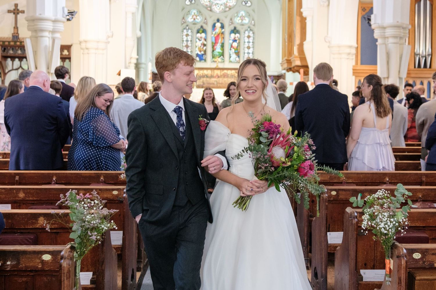 Traditional church wedding ceremony with couple smiling in aisle, decorated with white floral arrangements on pews.