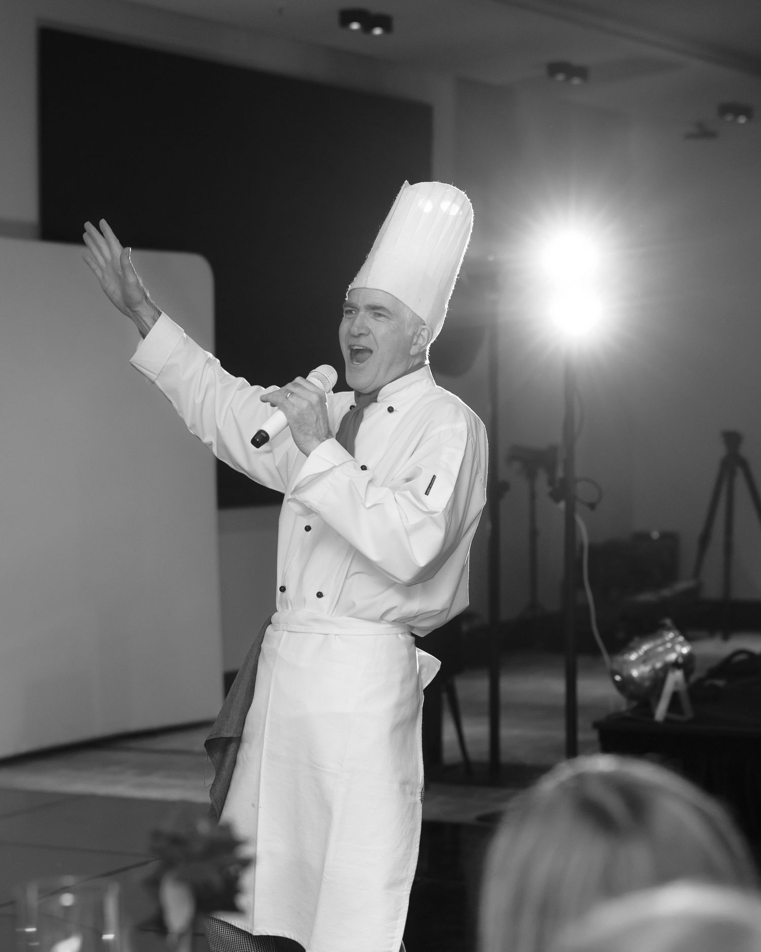 Chef in white uniform gestures enthusiastically during a presentation in a brightly lit room.