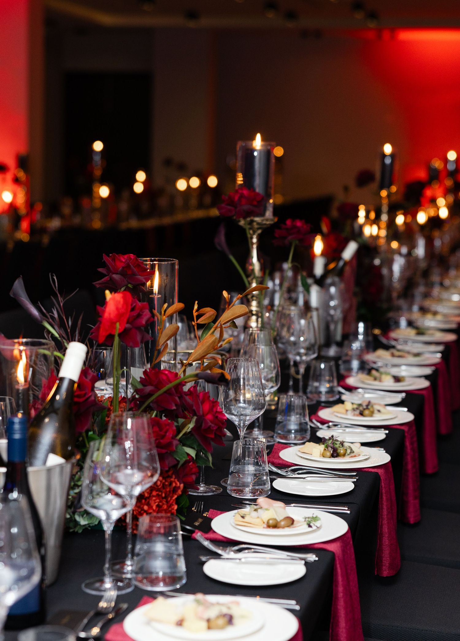 Elegant dinner table setting with red roses, candles and white plates against dark tablecloth in moody lighting.