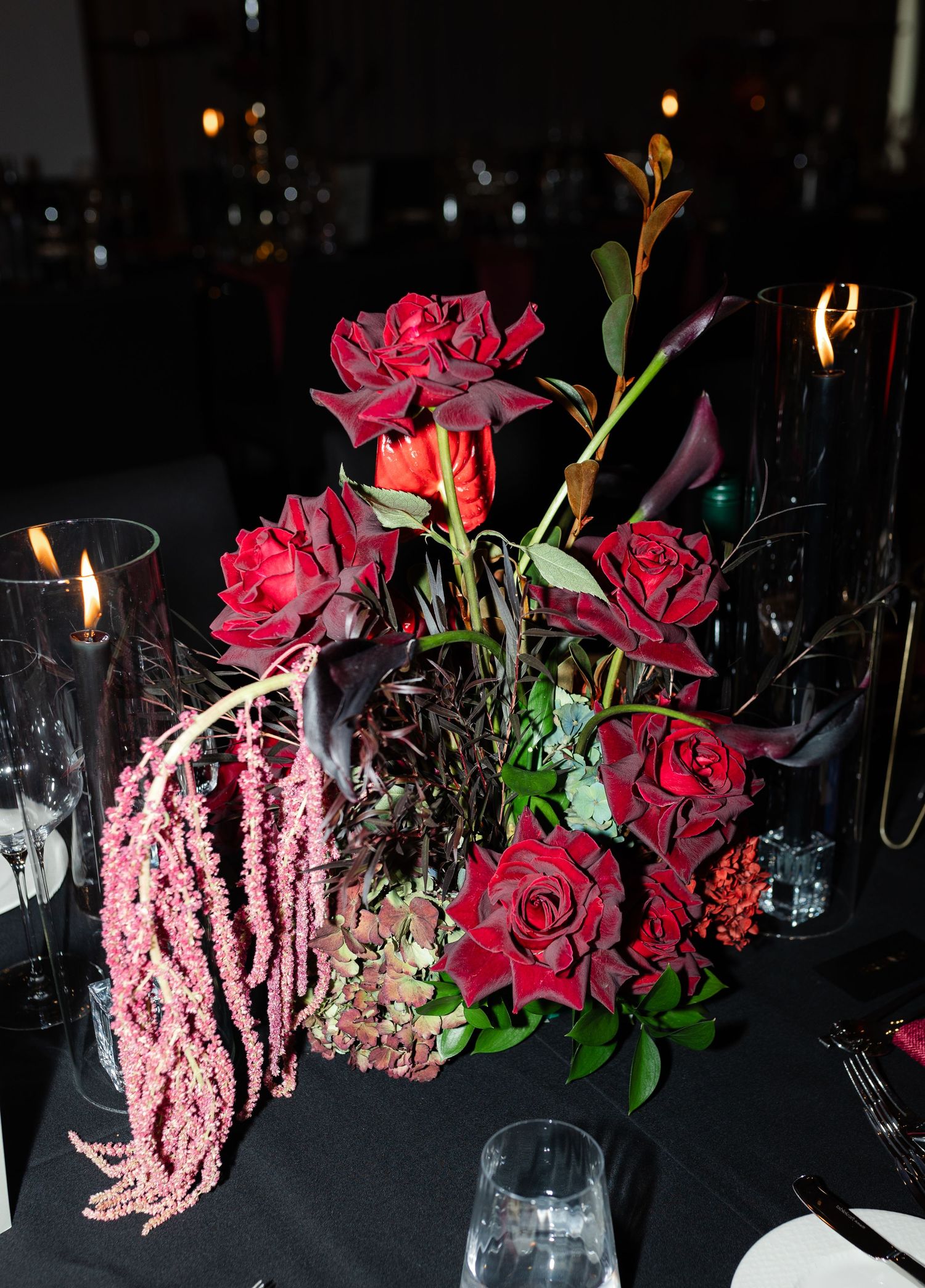 Elegant floral arrangement with deep red roses and pink amaranthus on dark table with candlelight.