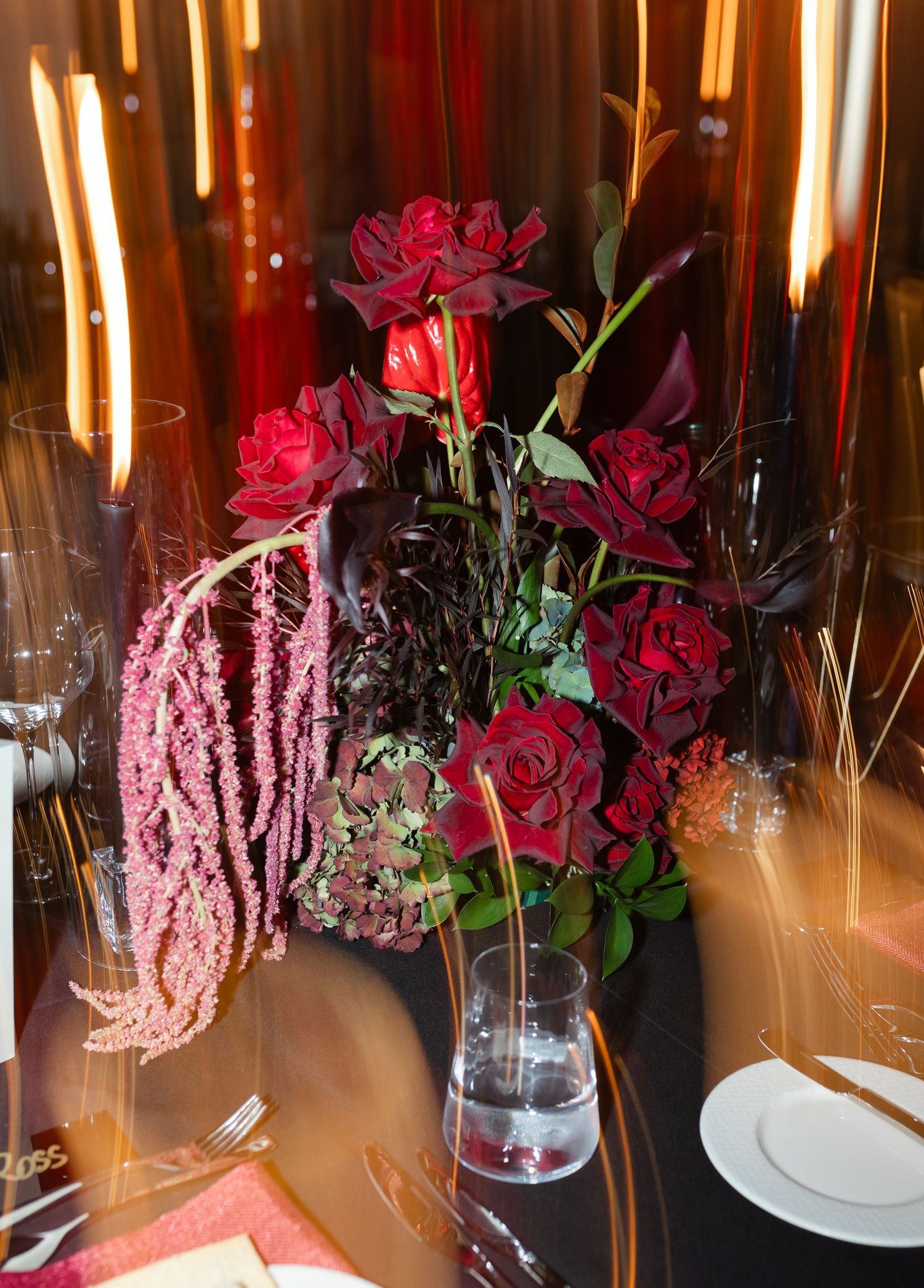 Elegant floral centerpiece with deep red roses and pink amaranthus in a glass vase against warm lighting and red drapery.