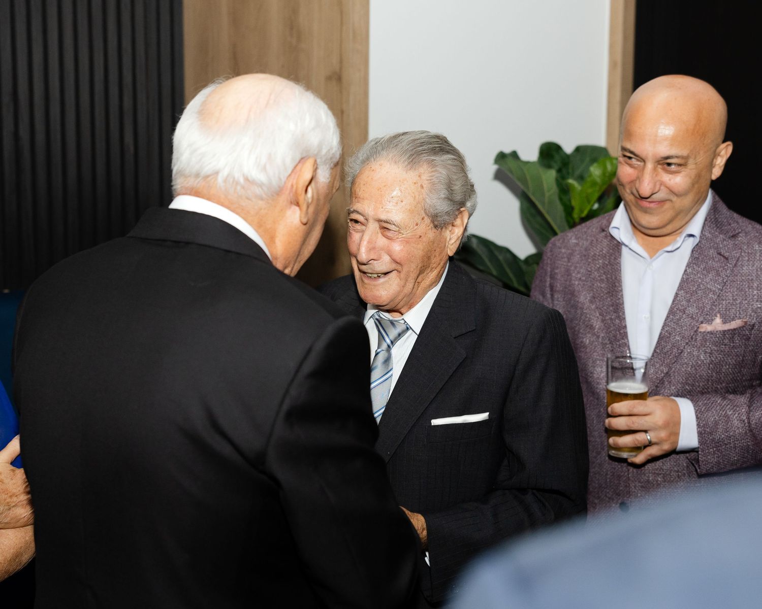 Three well-dressed men conversing and smiling at an indoor social gathering while holding drinks.
